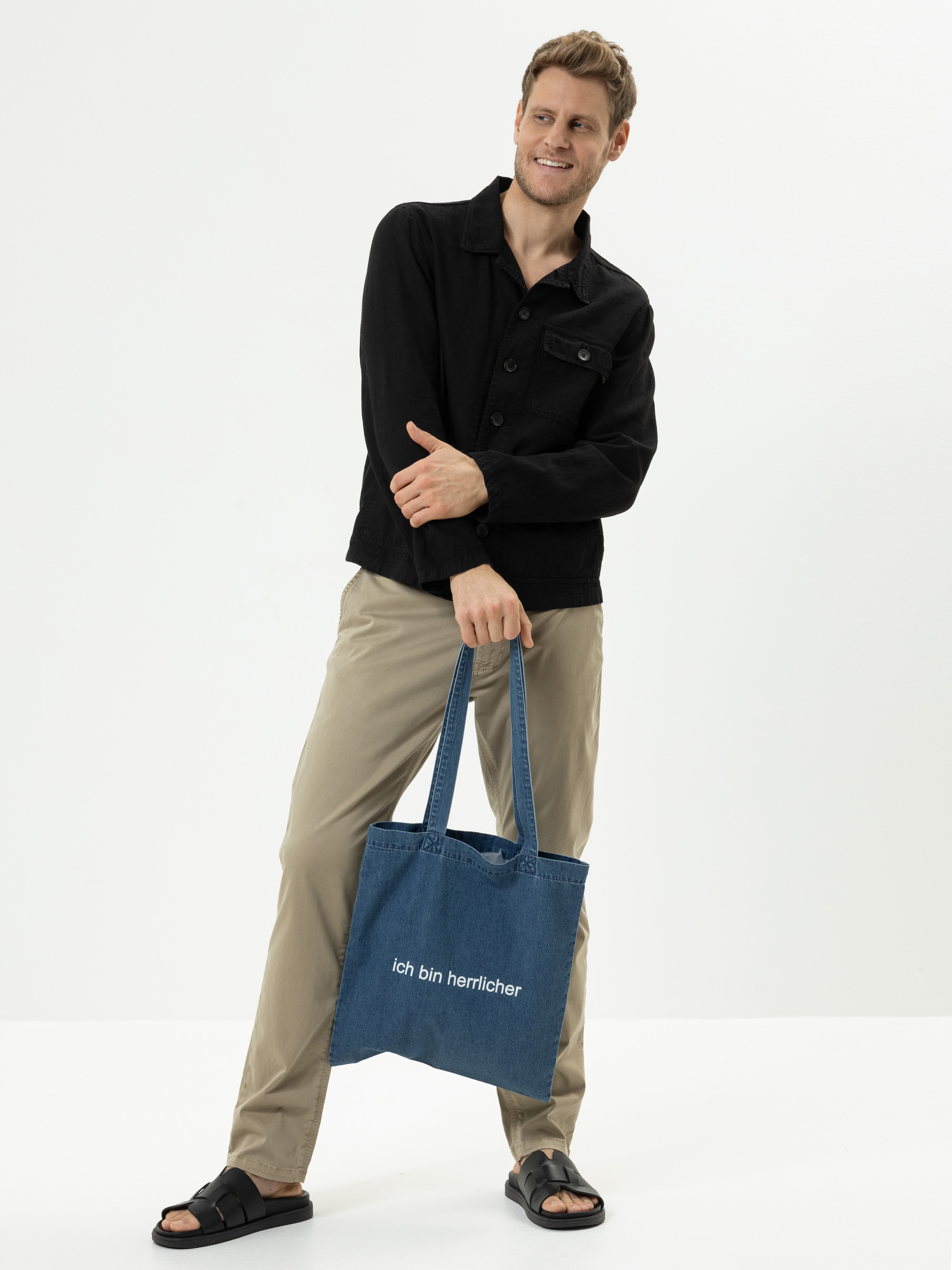 Ein Mann in schwarzer Jacke, beiger Hose und schwarzen Sandalen steht lächelnd da und hält eine blaue Tragetasche aus Jeansstoff mit der Aufschrift Ich bin herrlicher vor einem schlichten weißen Hintergrund.