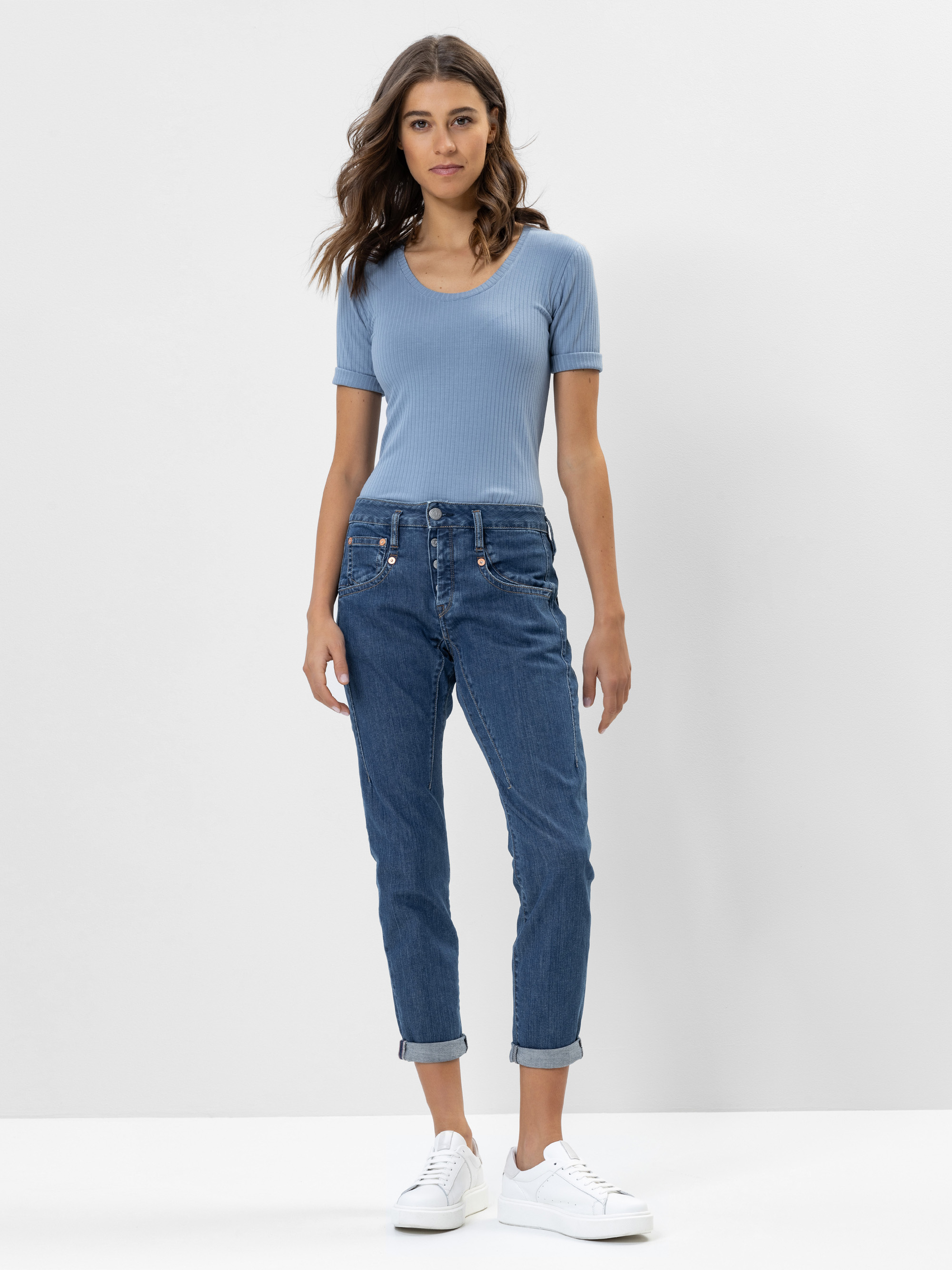 Eine Frau mit gewelltem braunem Haar steht vor einem schlichten weißen Hintergrund und trägt ein hellblaues, kurzärmeliges, geripptes Oberteil, hochgekrempelte Bluejeans und weiße Turnschuhe. Sie blickt mit neutralem Gesichtsausdruck nach vorne.