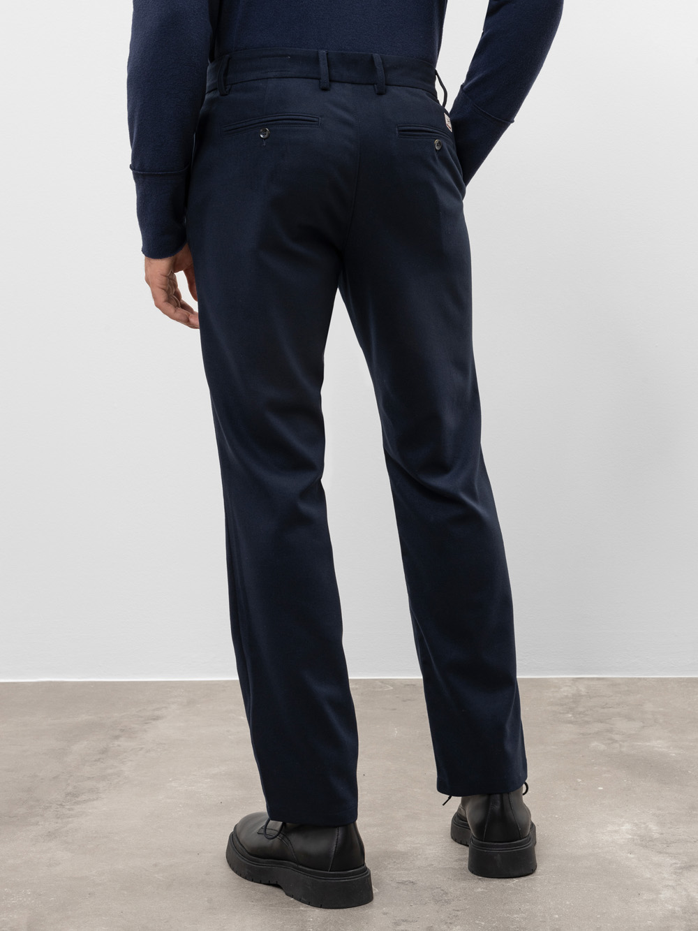 Eine Person, die eine dunkle marineblaue Anzughose, ein passendes marineblaues langärmeliges Hemd und schwarze Schuhe trägt, steht mit dem Gesicht zur Kamera auf einem Betonboden mit einem einfachen weißen Wandhintergrund.