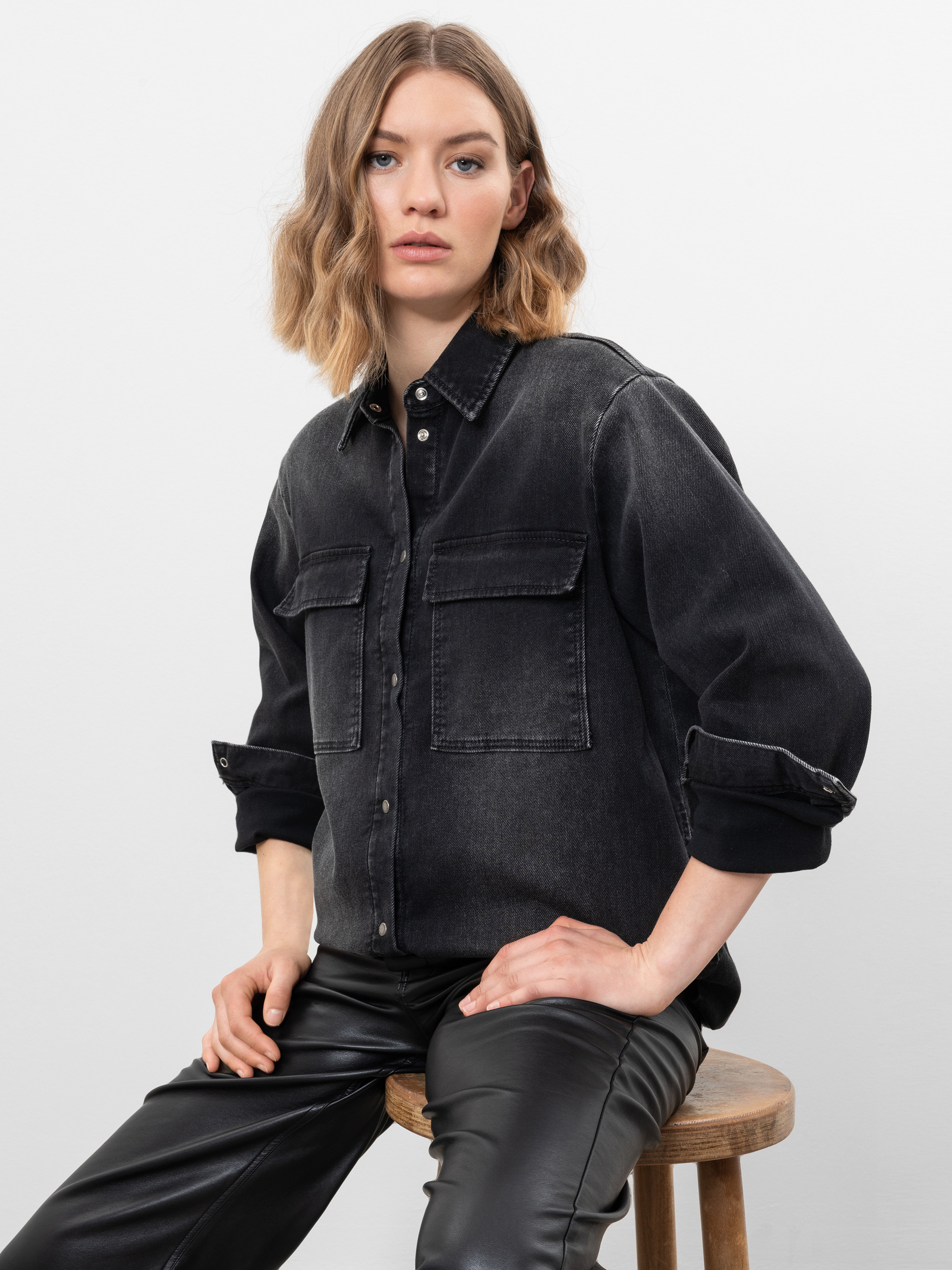 Eine Frau mit gewelltem, schulterlangem blondem Haar sitzt auf einem Holzschemel und trägt ein schwarzes Jeanshemd mit großen Vordertaschen und eine schwarze Lederhose vor einem schlichten weißen Hintergrund.