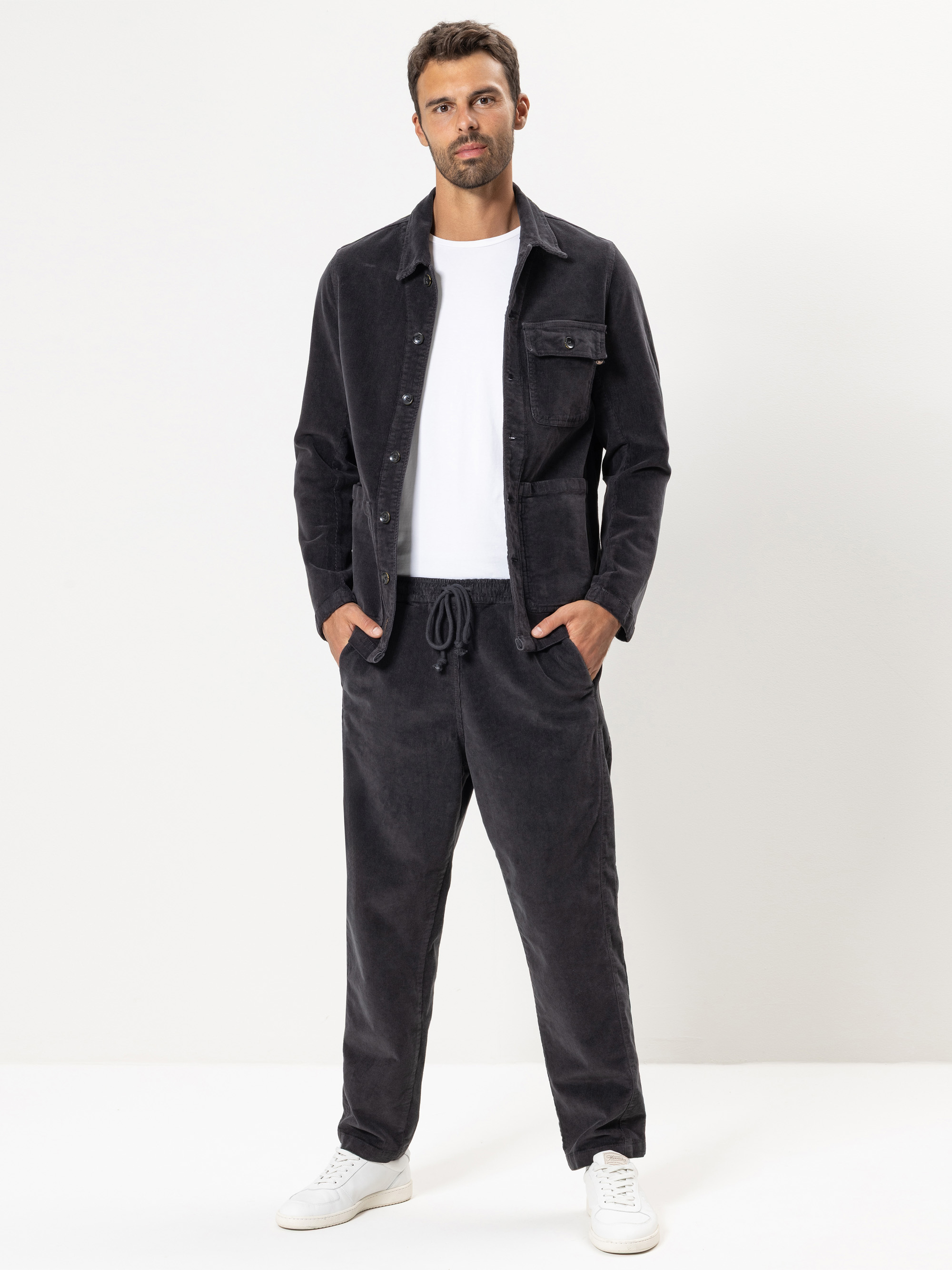 Ein Mann steht vor einem schlichten weißen Hintergrund, trägt eine schwarze Jacke mit Knöpfen, eine dazu passende schwarze Hose mit Kordelzug, ein weißes T-Shirt und weiße Turnschuhe, die Hände in den Taschen.