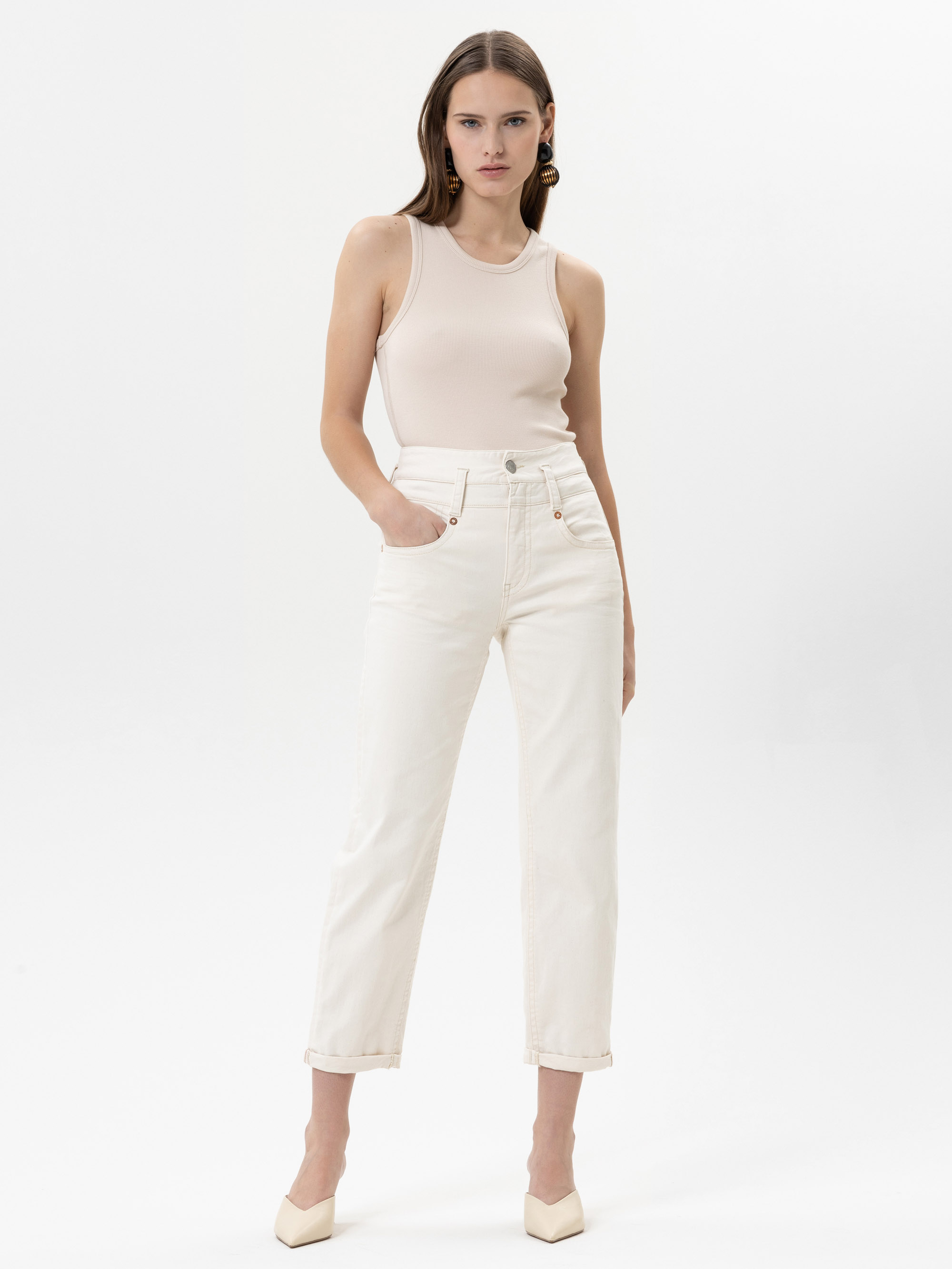 Eine Frau steht vor einem weißen Hintergrund, trägt ein ärmelloses, beigefarbenes Oberteil, weiße Jeans mit hoher Taille und cremefarbene Absätze, die Hände in den Taschen.