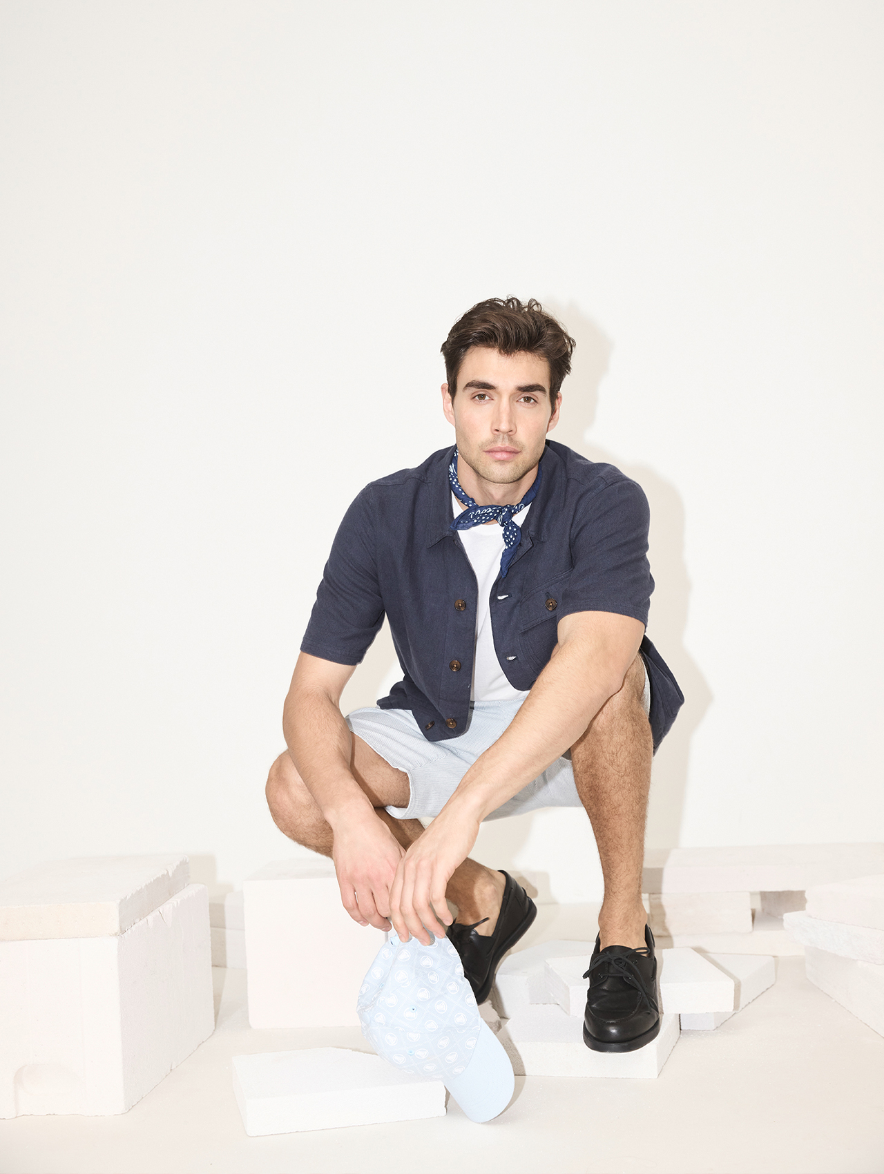 Ein Mann mit dunklem Haar, der ein marineblaues kurzärmeliges Hemd, ein weißes T-Shirt, helle Shorts und schwarze Schuhe trägt, kniet auf einem Knie und hält eine hellblaue Mütze mit weißen Mustern vor einem weißen Hintergrund mit weißen Blöcken.