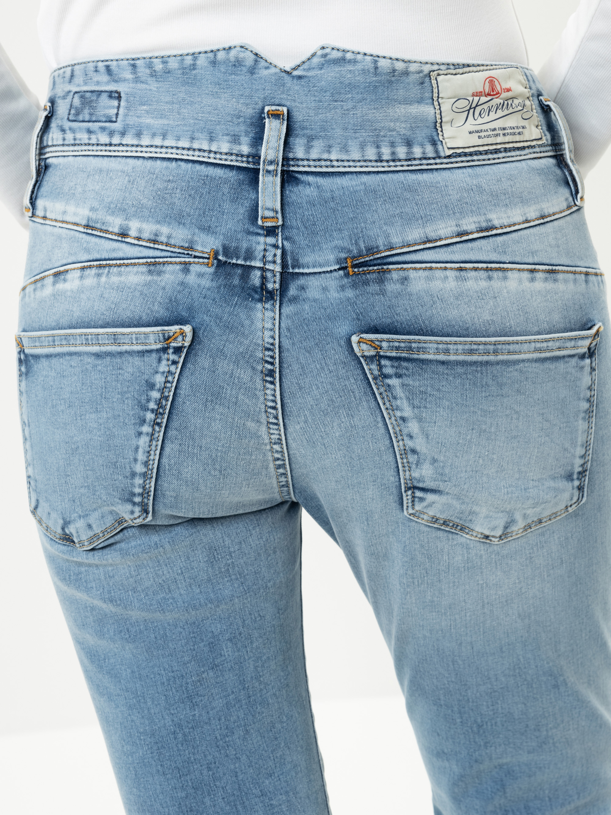Nahaufnahme des Rückens einer Person, die eine hellblaue Jeans mit zwei Gesäßtaschen, einer kleinen Gürtelschlaufe und einem weißen Aufnäher am Bund trägt. Die Person trägt außerdem ein weißes Hemd, das in die Hose gesteckt ist.
