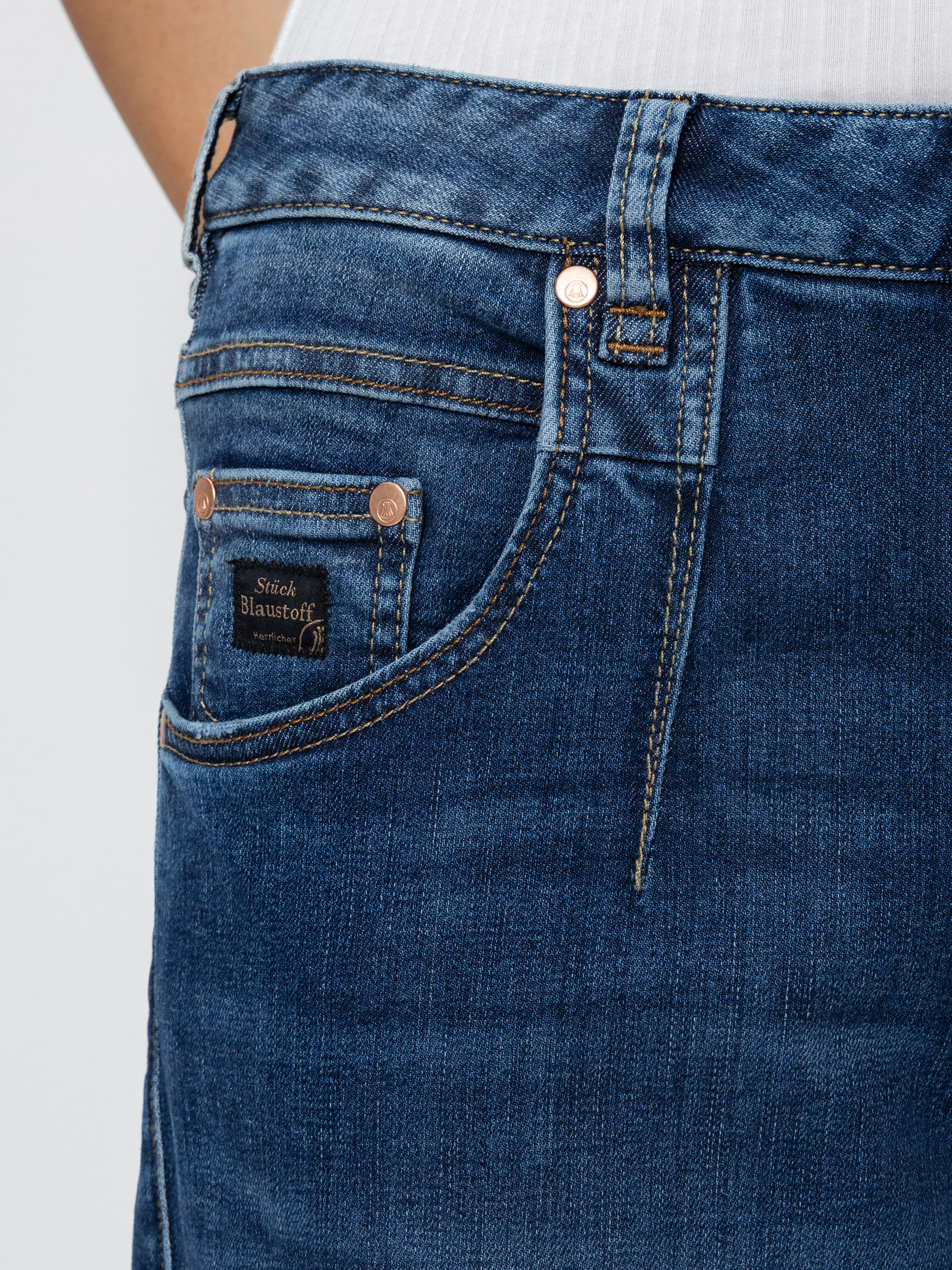 Nahaufnahme einer blauen Denim-Jeans mit Vordertasche, Münztasche und Kupfernieten, wobei ein Teil eines weißen gerippten Oberteils an der Taille sichtbar ist. Die Jeans hat sichtbare Nähte und ein kleines braunes Etikett auf der Münztasche.