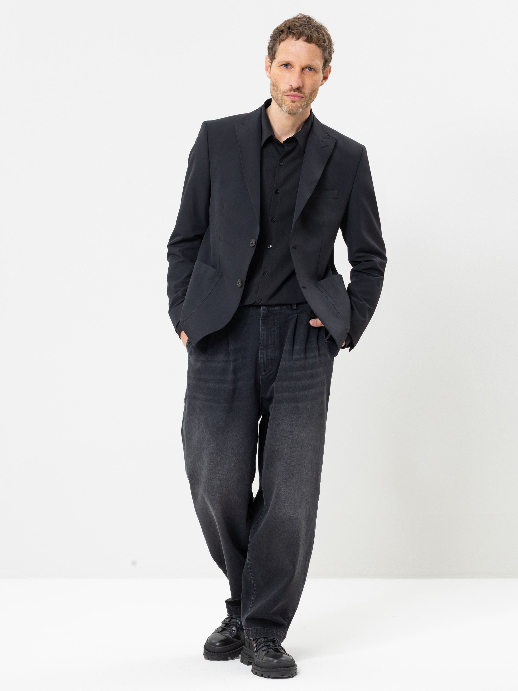 Ein Mann, der einen schwarzen Blazer, ein schwarzes Hemd, eine locker sitzende dunkle Jeans und schwarze Schuhe trägt, steht vor einem weißen Hintergrund, die Hände in den Taschen, und blickt in die Kamera.