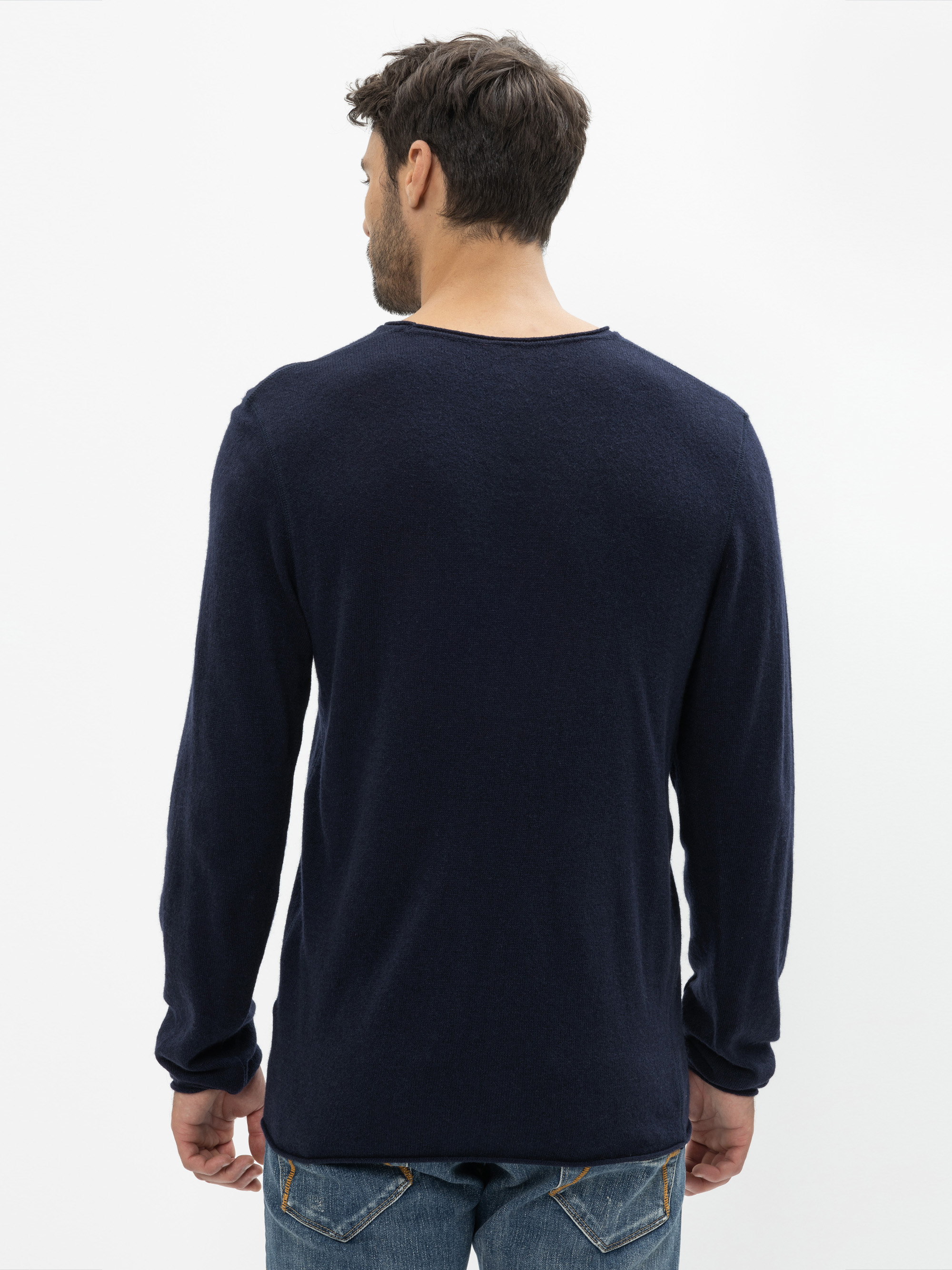 Ein Mann mit kurzen braunen Haaren, der einen dunkelblauen langärmeligen Pullover und eine blaue Jeans trägt, steht mit dem Gesicht zur Kamera vor einem schlichten weißen Hintergrund.