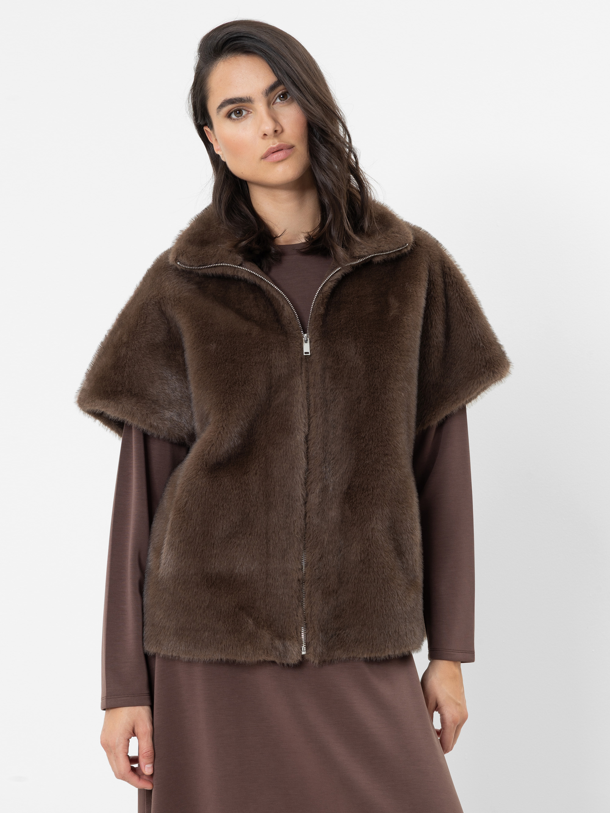 Eine Person, die eine braune Kunstfellweste mit Reißverschluss über einem braunen Langarmkleid trägt, steht vor einem schlichten weißen Hintergrund und blickt mit ausdruckslosem Gesichtsausdruck in die Kamera.