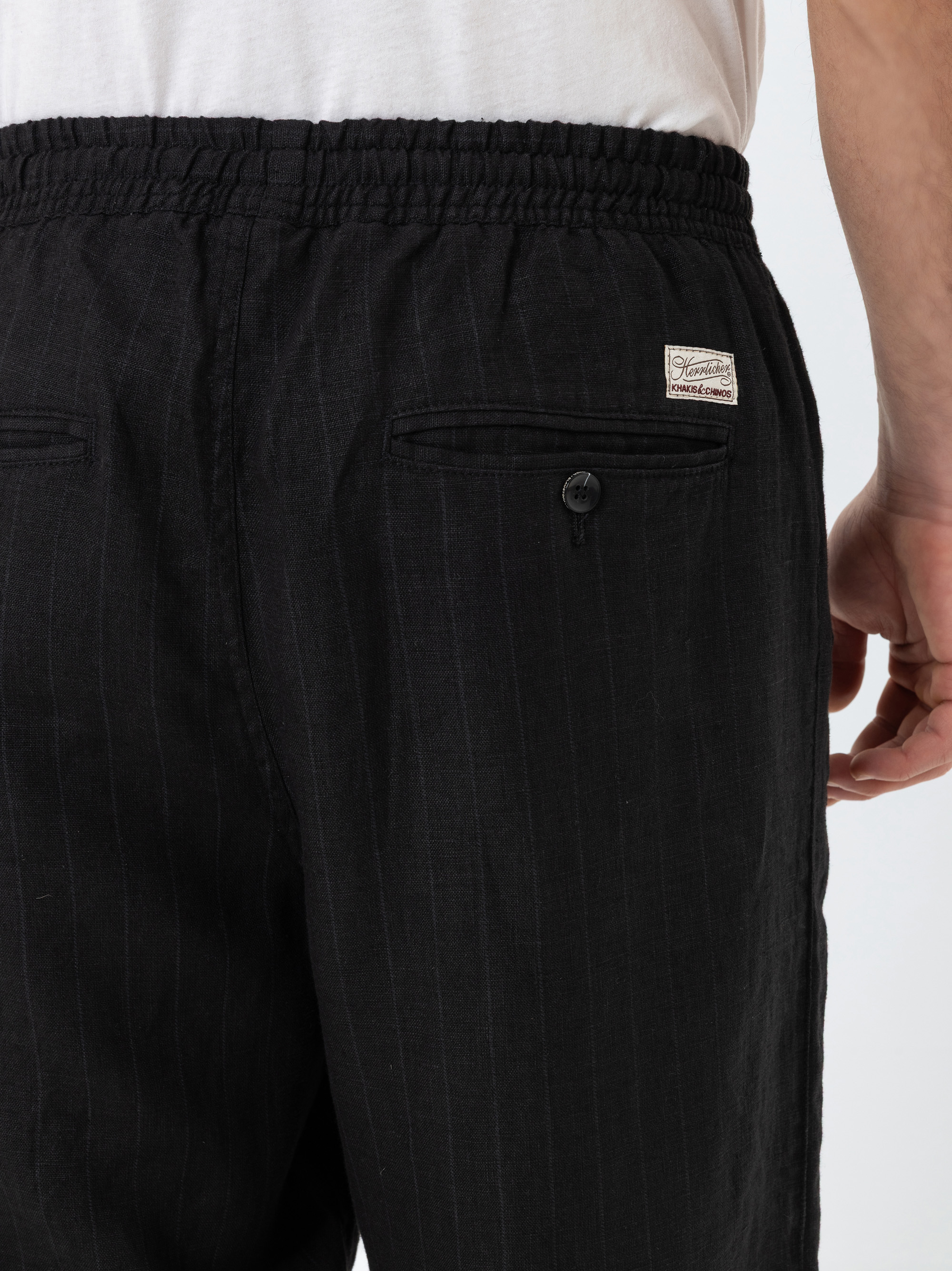 Eine Person, die eine schwarze Nadelstreifenhose mit einem elastischen Bund und einer Gesäßtasche mit Knopf trägt, wird von hinten gezeigt. Ein Teil eines weißen T-Shirts und die Hand der Person sind zu sehen.