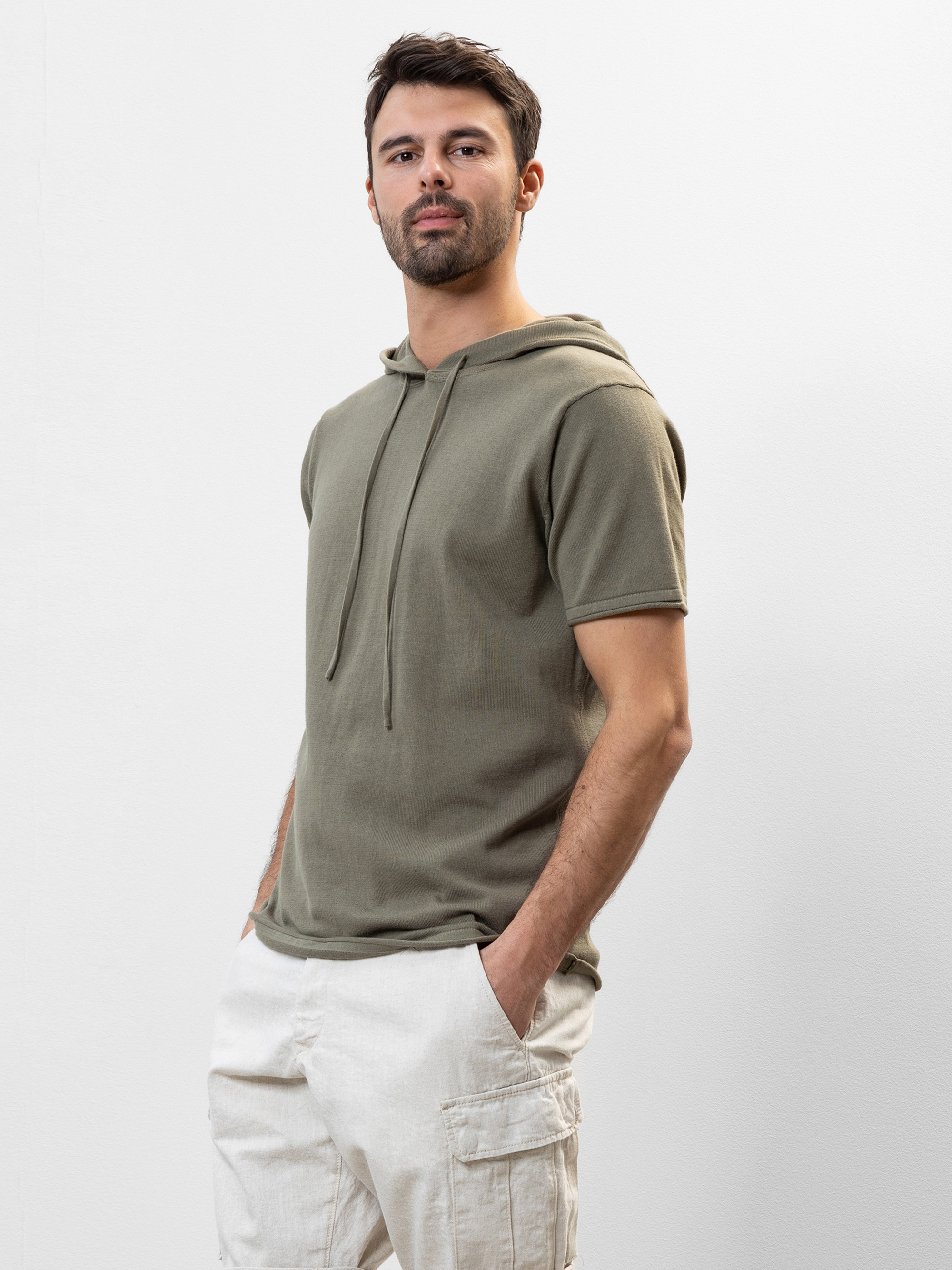 Ein Mann mit kurzen dunklen Haaren und einem Bart trägt einen olivgrünen, kurzärmeligen Kapuzenpullover und eine helle Cargohose. Er steht mit den Händen in den Taschen vor einem schlichten weißen Hintergrund.