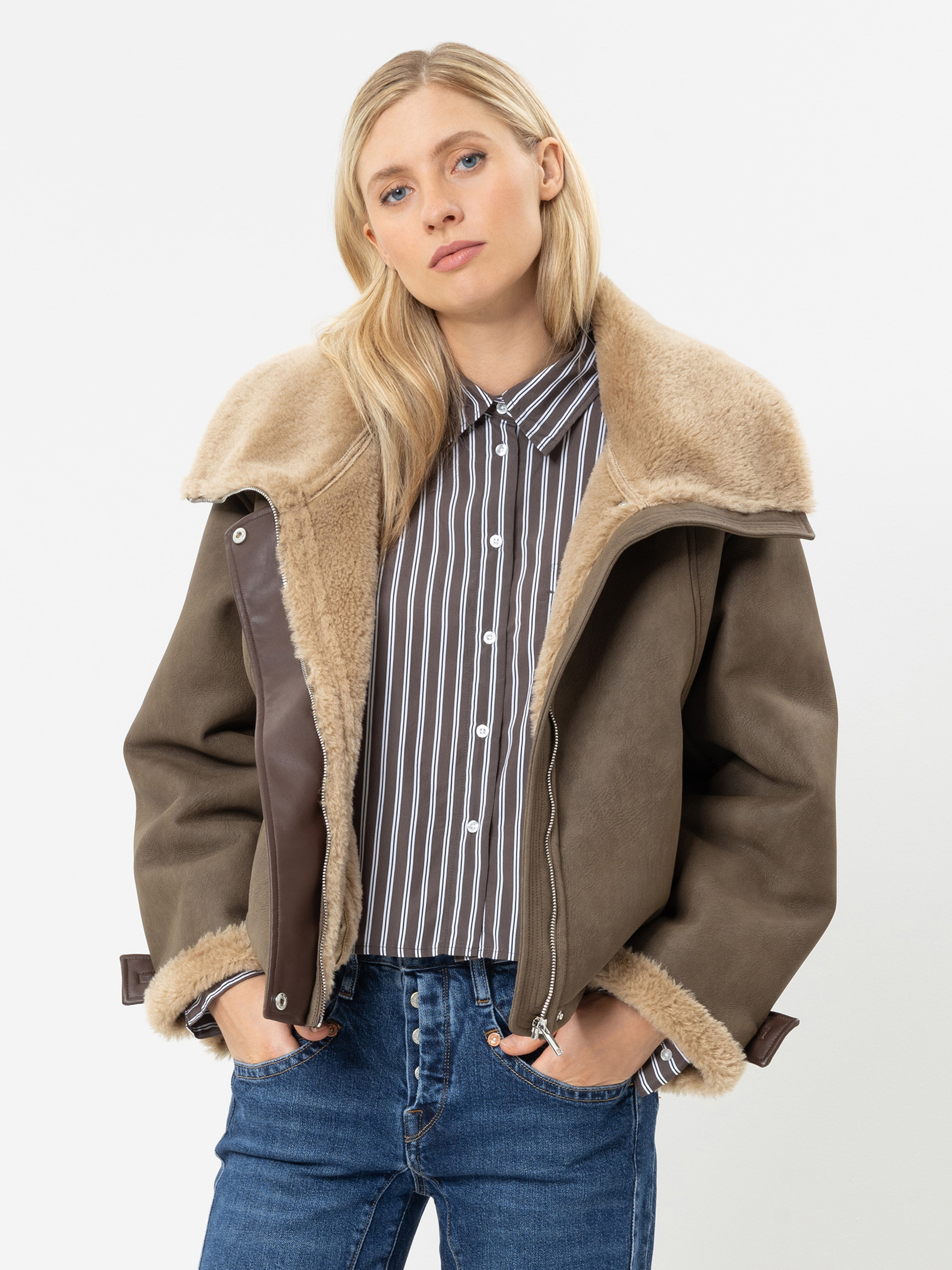 Eine Person mit glattem blondem Haar trägt eine braune Lammfelljacke über einem gestreiften Hemd und blauen Jeans und steht vor einem schlichten weißen Hintergrund.