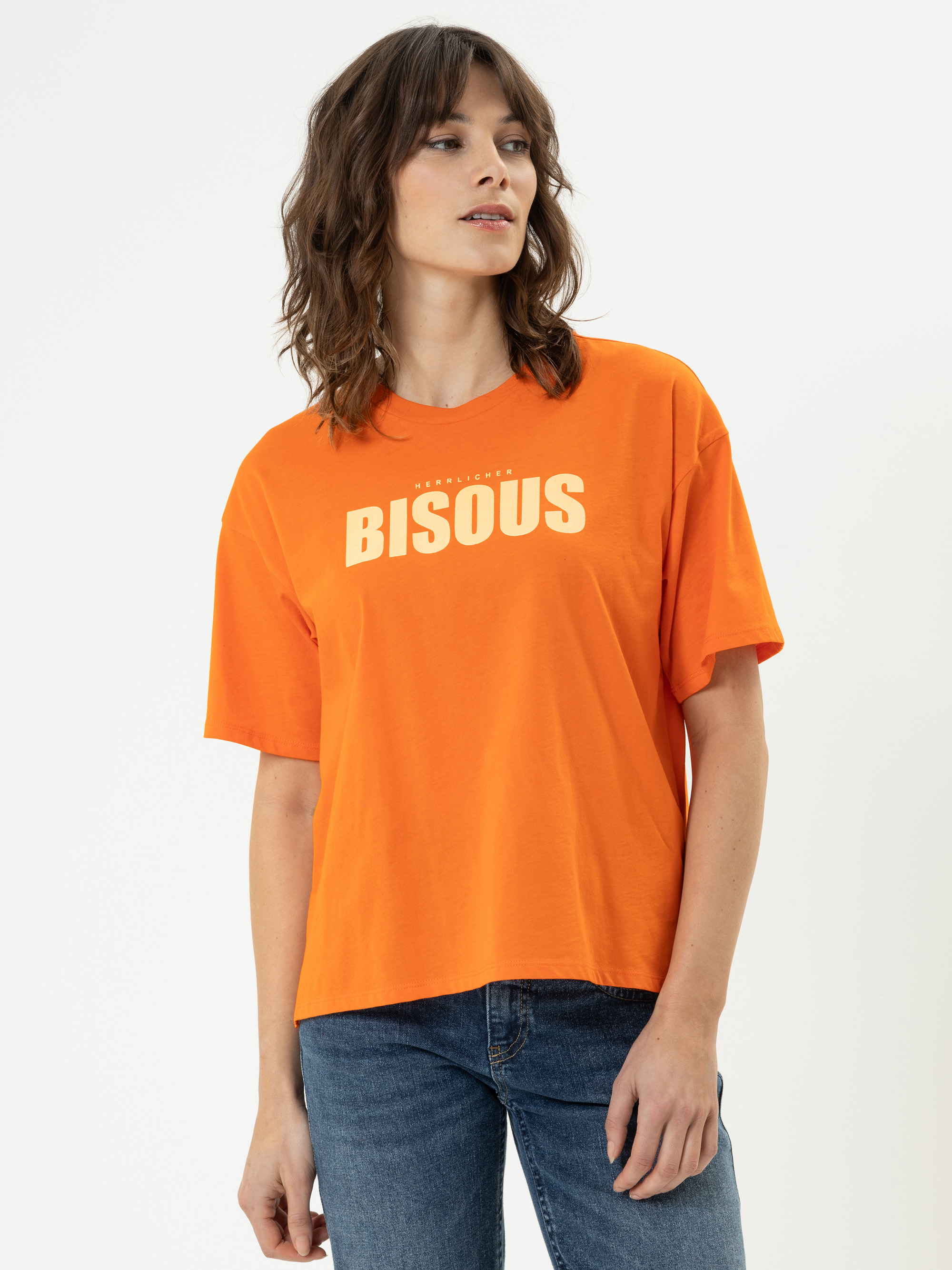 Eine Frau mit gewellten braunen Haaren trägt ein orangefarbenes T-Shirt mit dem weiß aufgedruckten Wort BISOUS auf der Brust, gepaart mit einer blauen Jeans, vor einem schlichten weißen Hintergrund.