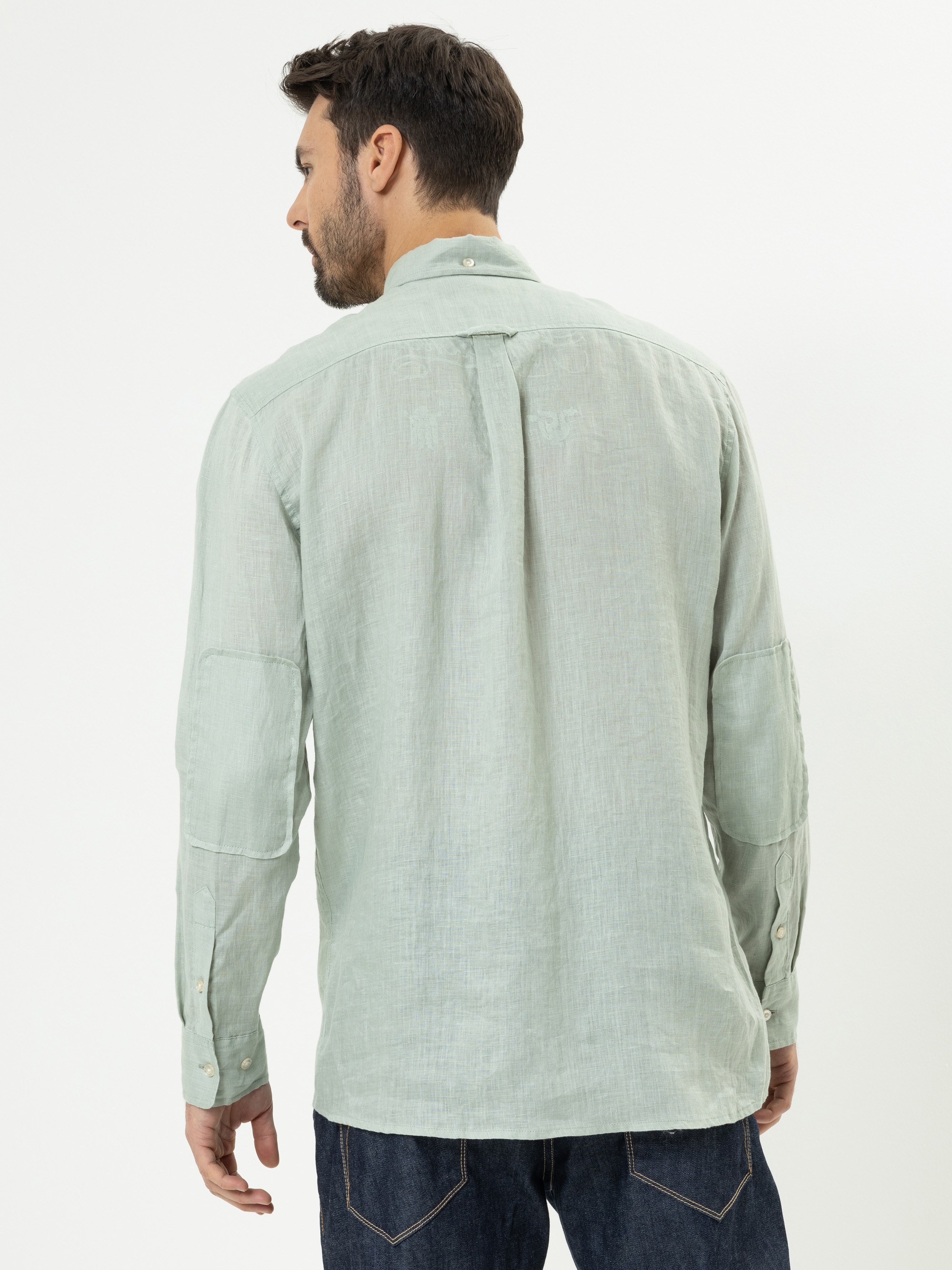 Ein Mann mit kurzen dunklen Haaren und Bart ist von hinten zu sehen. Er trägt ein hellgrünes, langärmeliges Button-up-Hemd und dunkelblaue Jeans vor einem schlichten weißen Hintergrund.