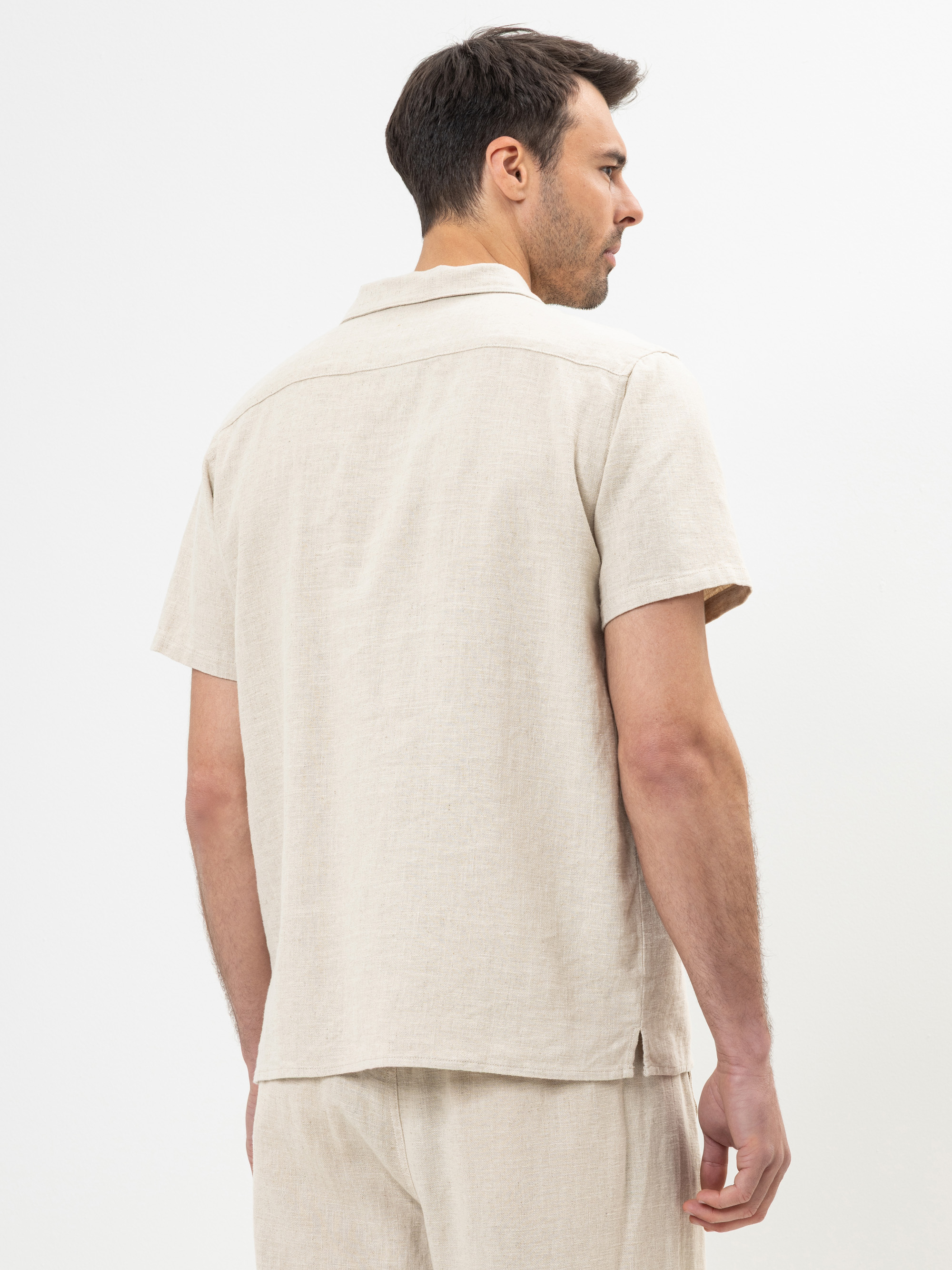 Ein Mann steht mit dem Rücken zur Kamera, trägt ein hellbeiges, kurzärmeliges Leinenhemd und passende Shorts vor einem schlichten weißen Hintergrund.