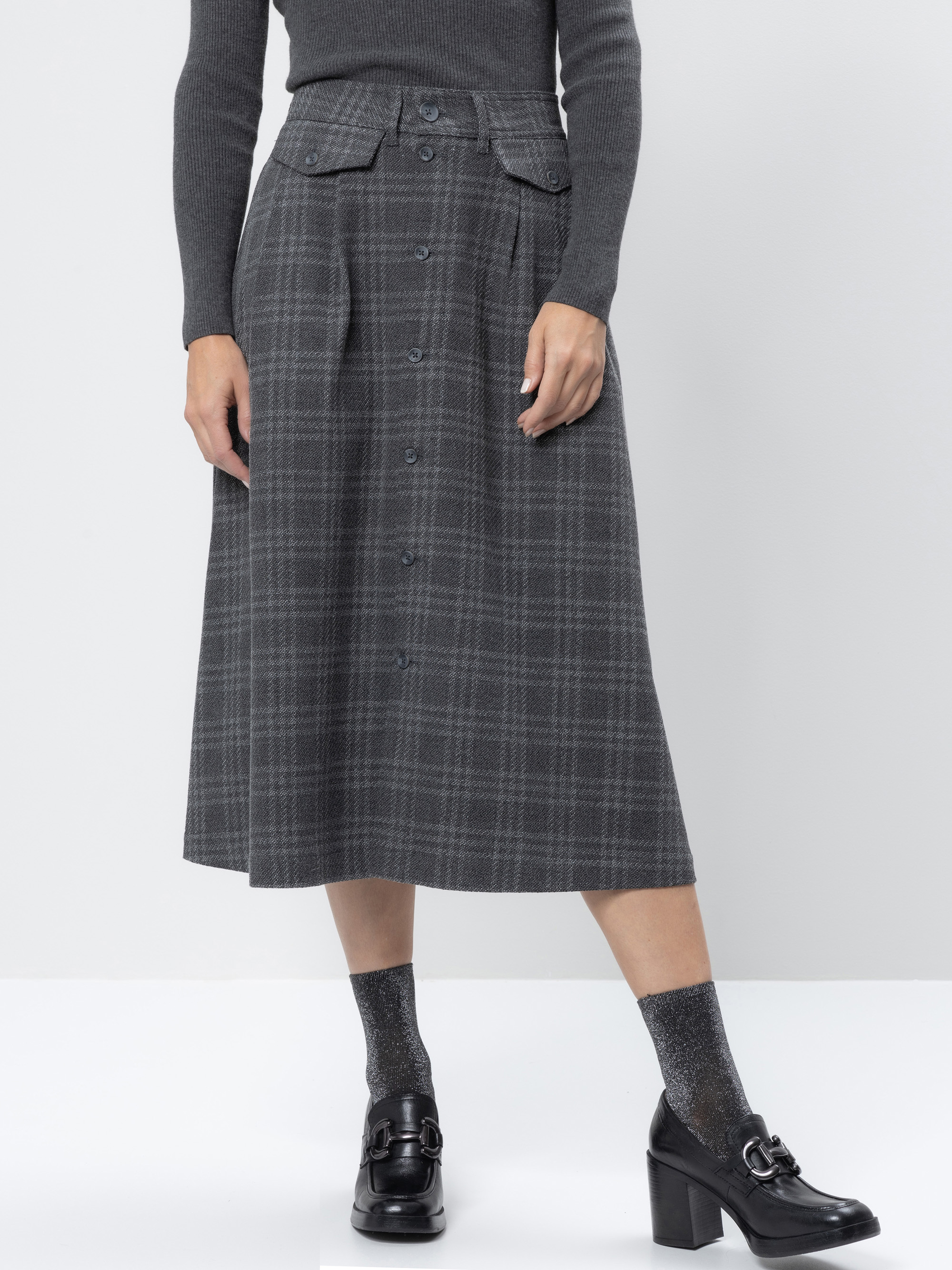 Eine Person, die einen hochtaillierten, geknöpften, grau karierten Midirock mit Vordertaschen, ein geripptes dunkelgraues Oberteil, glitzernde graue Socken und schwarze Halbschuhe mit Absatz trägt, steht vor einem schlichten hellen Hintergrund.