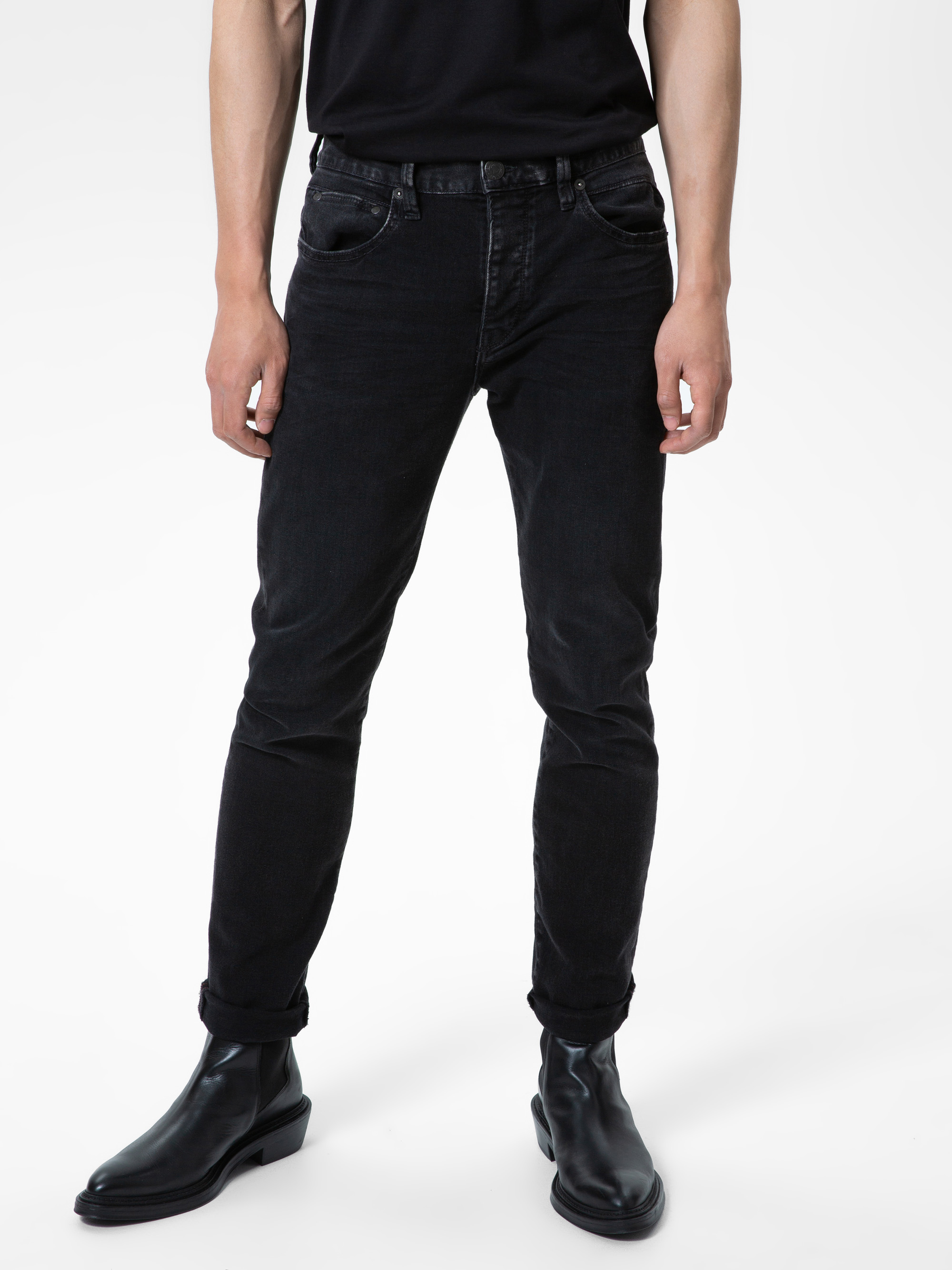 Eine Person, die eine schwarze Jeans, ein schwarzes T-Shirt und schwarze Lederstiefel trägt und auf einem weißen Hintergrund steht. Das Bild zeigt die Person nur von den Schultern abwärts.