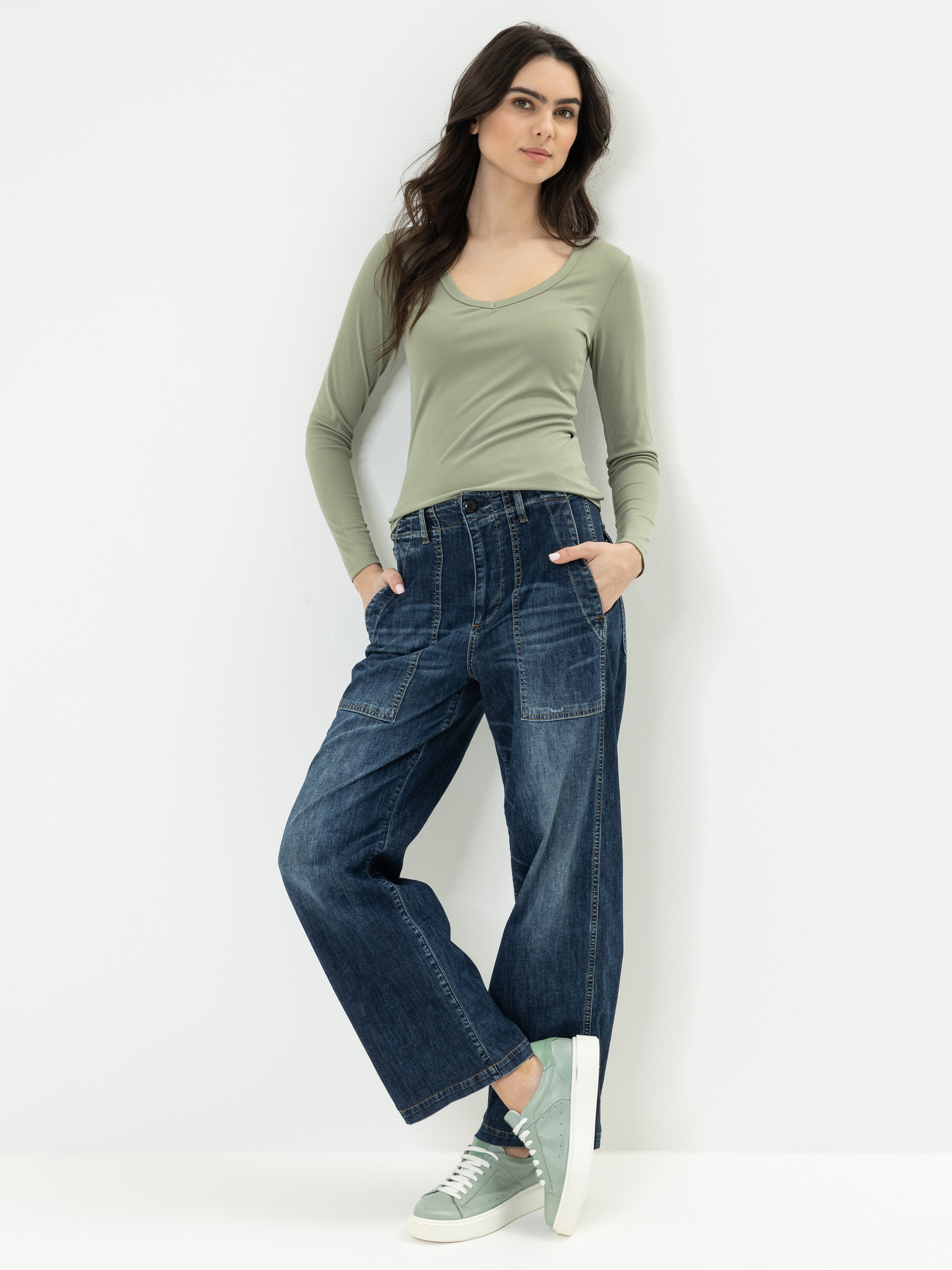 Eine Frau steht vor einem weißen Hintergrund und trägt ein hellgrünes, langärmeliges Oberteil, weit geschnittene blaue Jeans mit sichtbaren Nähten und hellgrüne Turnschuhe. Sie hat die Hände in den Taschen und trägt langes dunkles Haar.