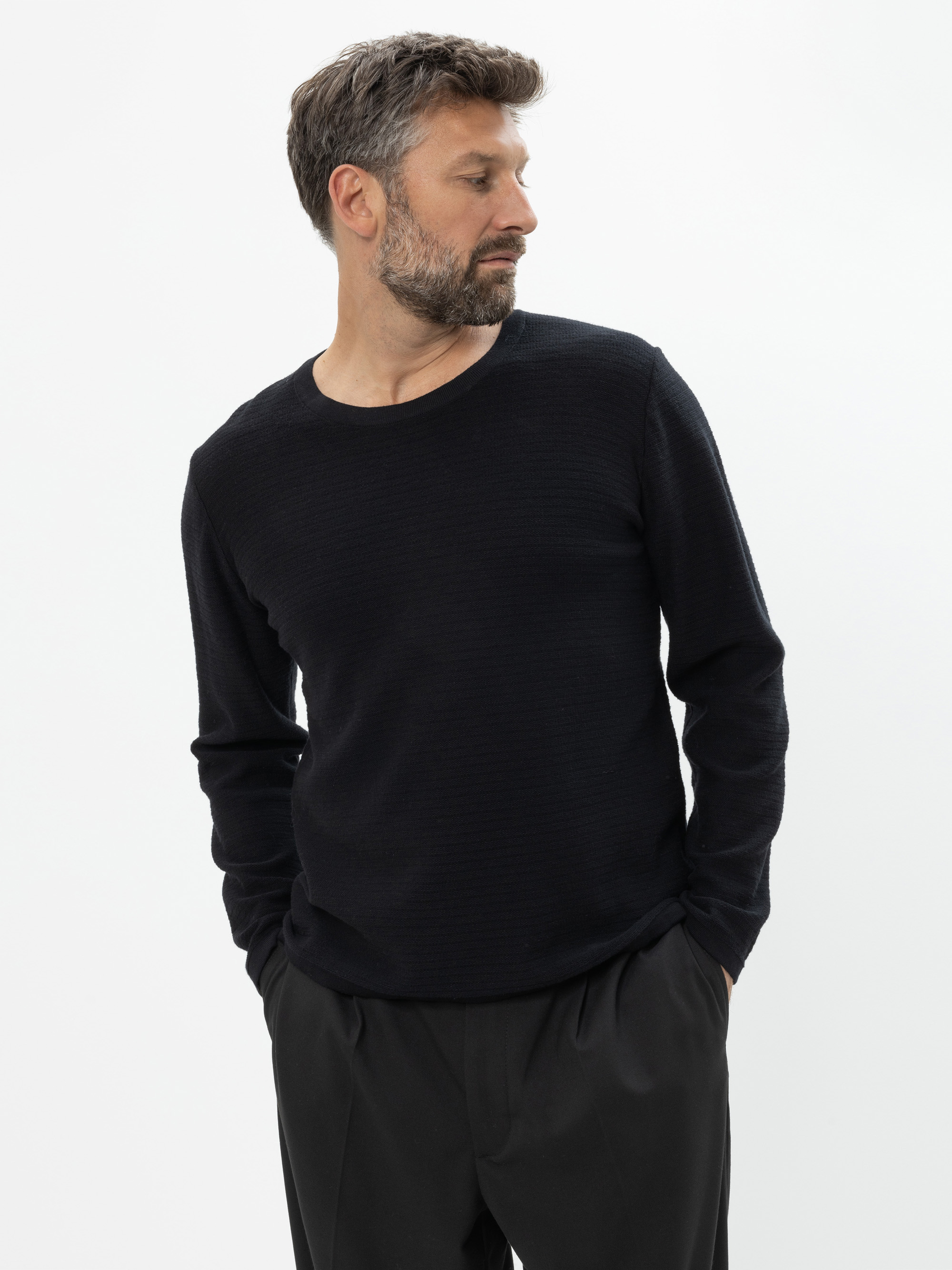 Ein Mann mit kurzen braunen Haaren und Bart, der ein schwarzes langärmeliges Hemd und eine schwarze Hose trägt, steht mit den Händen in den Taschen und blickt nach links vor einem schlichten weißen Hintergrund.