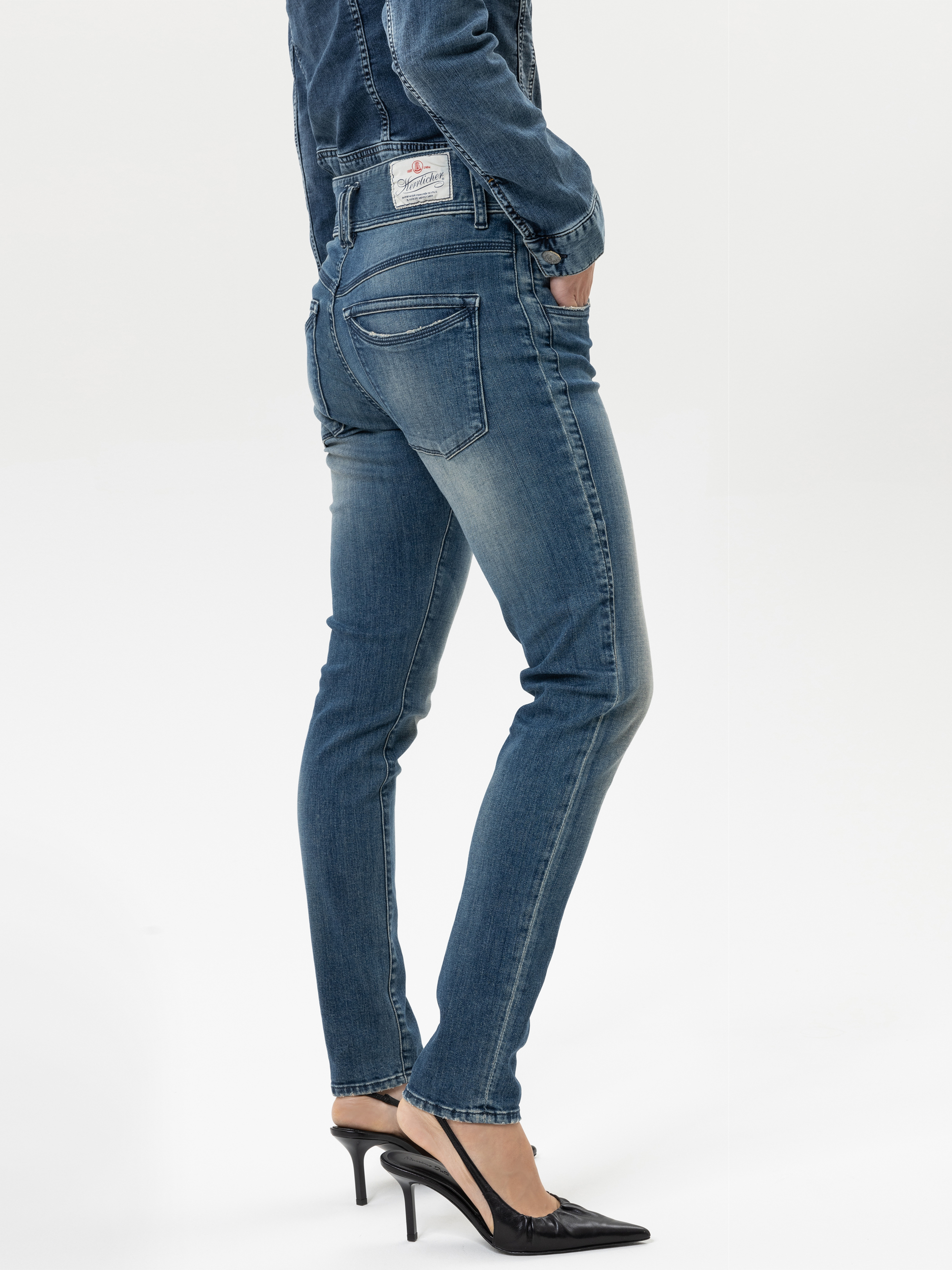 Eine Person in Bluejeans und schwarzen Stöckelschuhen steht mit einem leicht angewinkelten Bein, die Hände in den Gesäßtaschen und einer Jeansjacke, die teilweise sichtbar ist; der Hintergrund ist schlicht weiß.