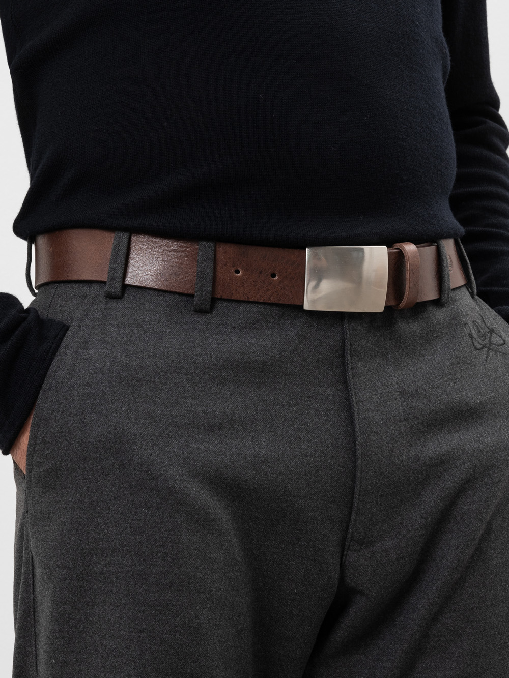 Eine Person, die ein schwarzes langärmeliges Hemd, eine graue Hose und einen braunen Ledergürtel mit einer großen silbernen Schnalle trägt und eine Hand in der Tasche hat.