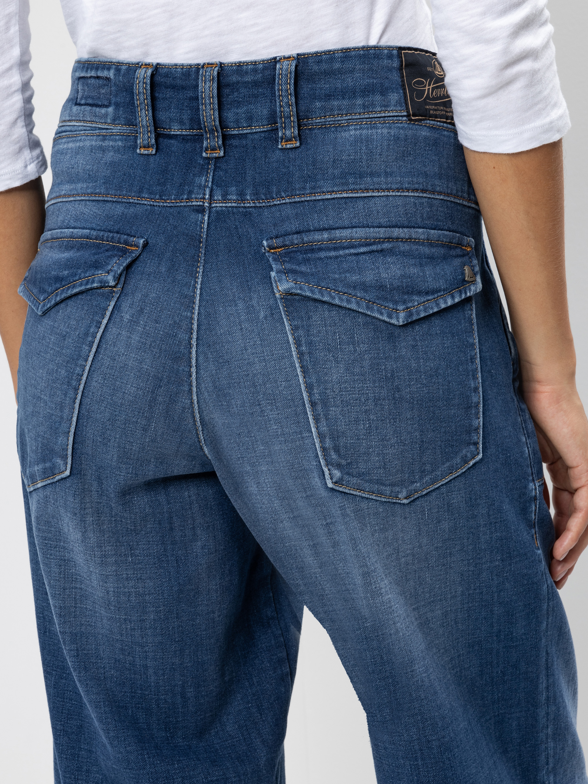 Nahaufnahme des Rückens einer Person, die eine blaue Jeans und ein weißes Hemd trägt. Man sieht den Bund, die Gürtelschlaufen und zwei Gesäßtaschen mit Klappen.