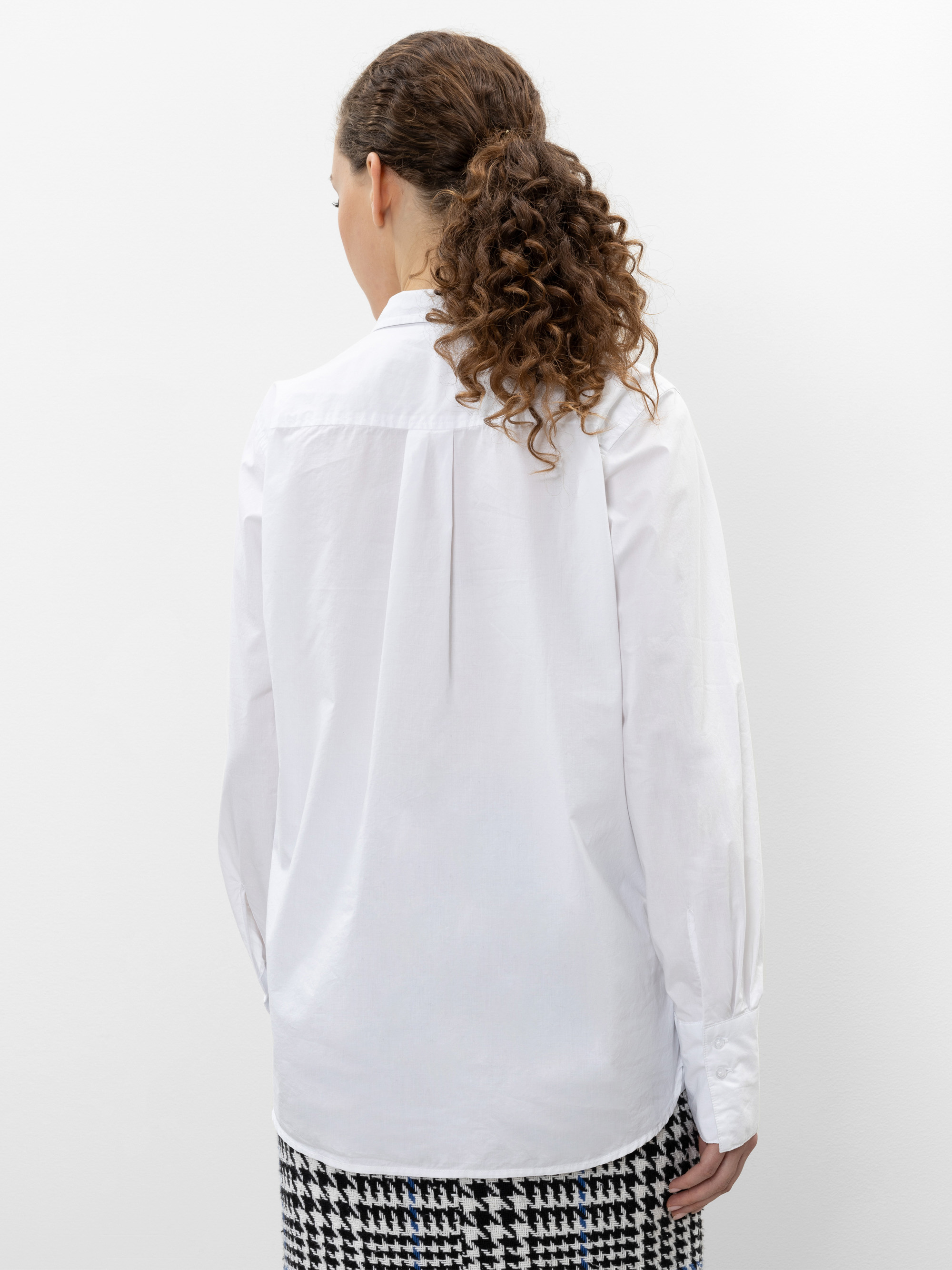 Eine Frau mit lockigem braunem Haar in einem Pferdeschwanz blickt von der Kamera weg und trägt ein weißes langärmeliges Hemd und einen schwarz-weißen Hahnentritt-Rock vor einem schlichten weißen Hintergrund.
