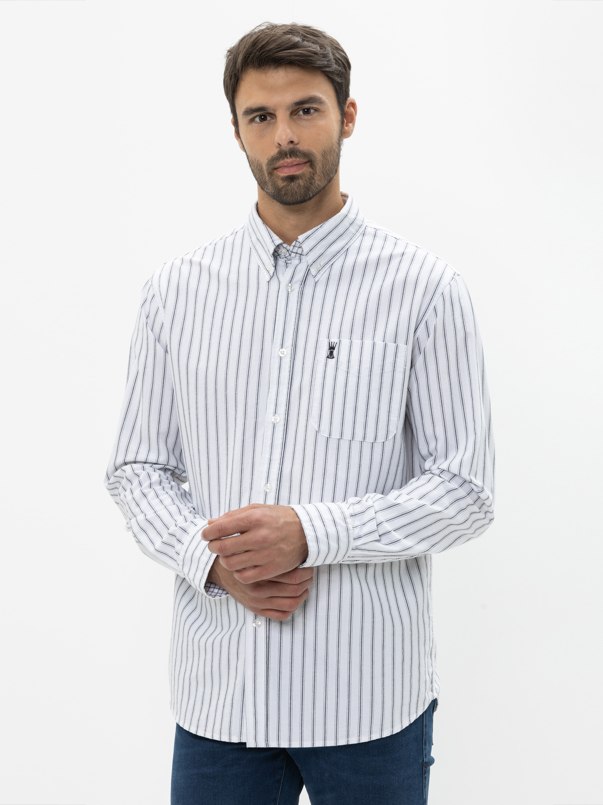 Ein Mann mit kurzen dunklen Haaren und Bart trägt ein weißes, geknöpftes Hemd mit schwarzen Längsstreifen und blaue Jeans vor einem schlichten weißen Hintergrund.