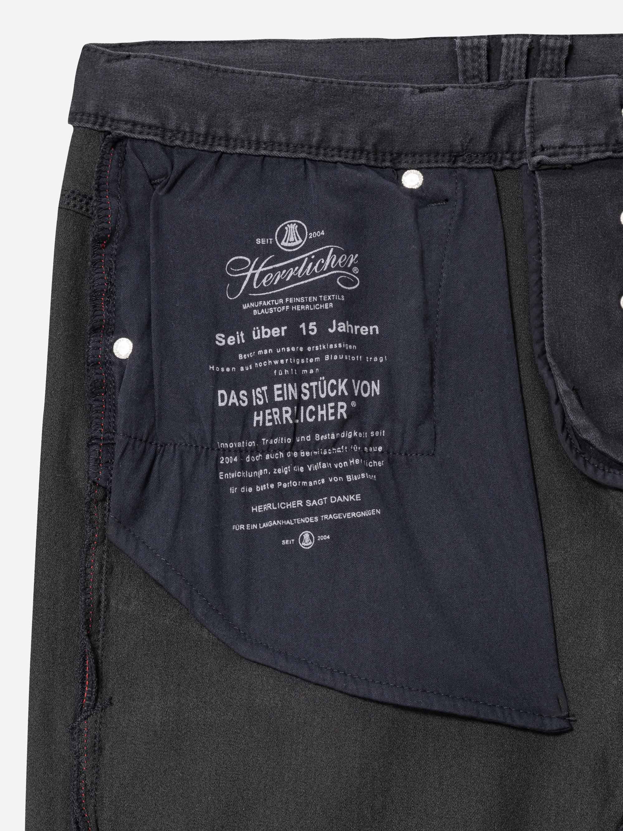 Nahaufnahme der Innentasche einer dunklen Jeans, die ein eingenähtes Stoffetikett mit weißem deutschen Text und Branding-Details von Herrlicher zeigt. Die Nähte und die Textur der Jeans sind sichtbar.