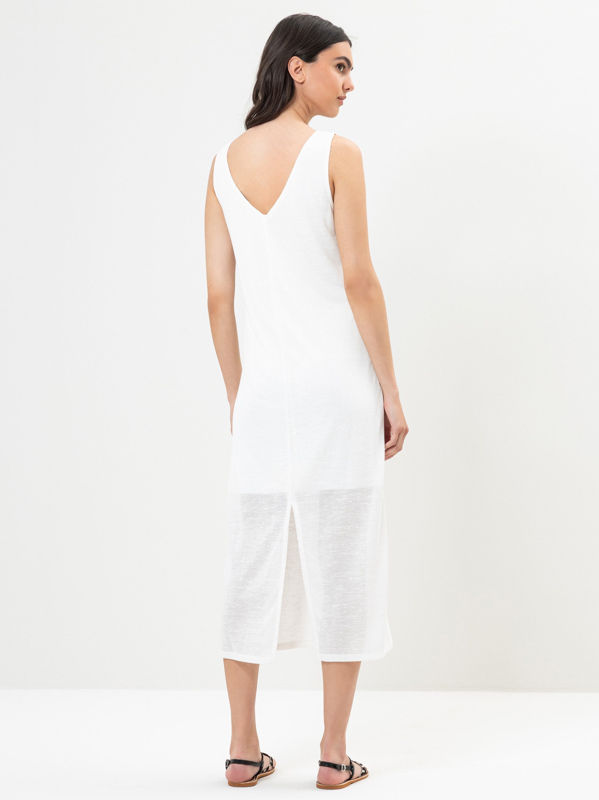 Eine Frau steht mit dem Gesicht zur Wand und trägt ein ärmelloses weißes Midikleid mit V-förmigem Rücken und einem Mittelschlitz, dazu schwarze flache Sandalen. Sie hat dunkles, gewelltes Haar und steht vor einem schlichten weißen Hintergrund.