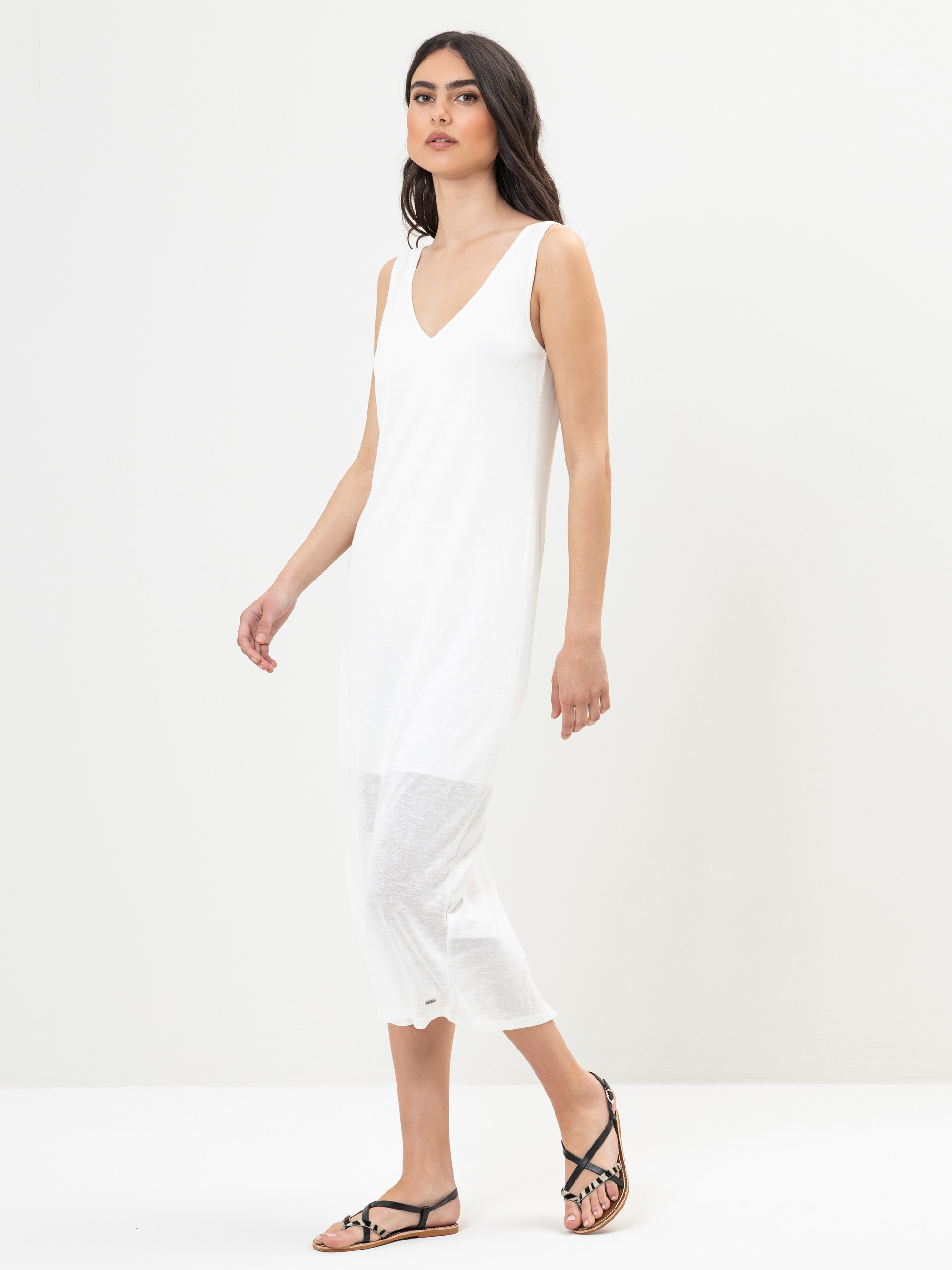 Eine Frau mit langen dunklen Haaren trägt ein ärmelloses, knöchellanges weißes Kleid mit V-Ausschnitt und schwarze Riemchensandalen und läuft vor einem schlichten weißen Hintergrund.