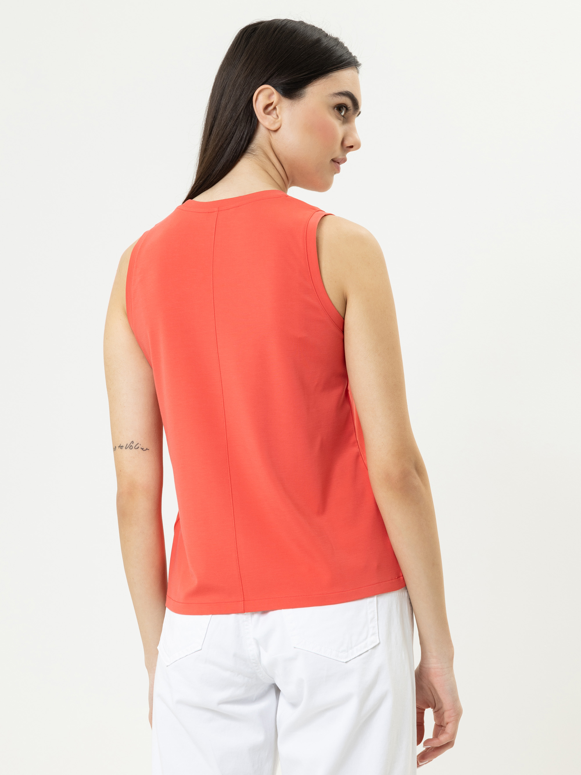 Eine Frau mit langen dunklen Haaren, die ein ärmelloses korallenrotes Oberteil und eine weiße Hose trägt, steht etwas abseits und zeigt die Rückseite ihres Outfits vor einem schlichten hellen Hintergrund. Auf ihrem linken Unterarm ist eine kleine Tät
