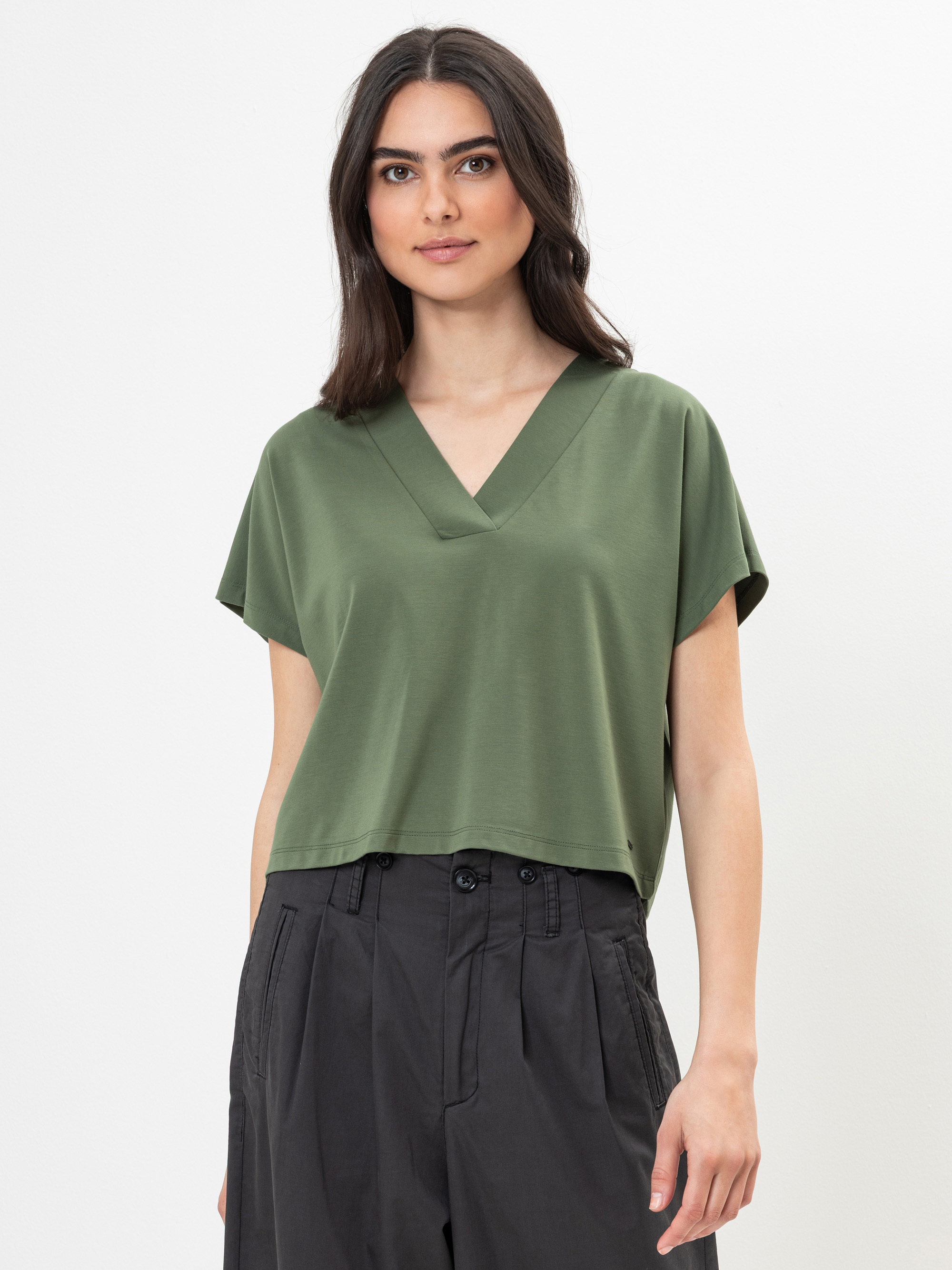 Eine Frau mit einem olivgrünen kurzärmeligen Oberteil mit V-Ausschnitt und einer hochgeschlossenen kohlegrauen Hose steht vor einem schlichten weißen Hintergrund. Sie hat langes dunkles Haar und einen neutralen Gesichtsausdruck.