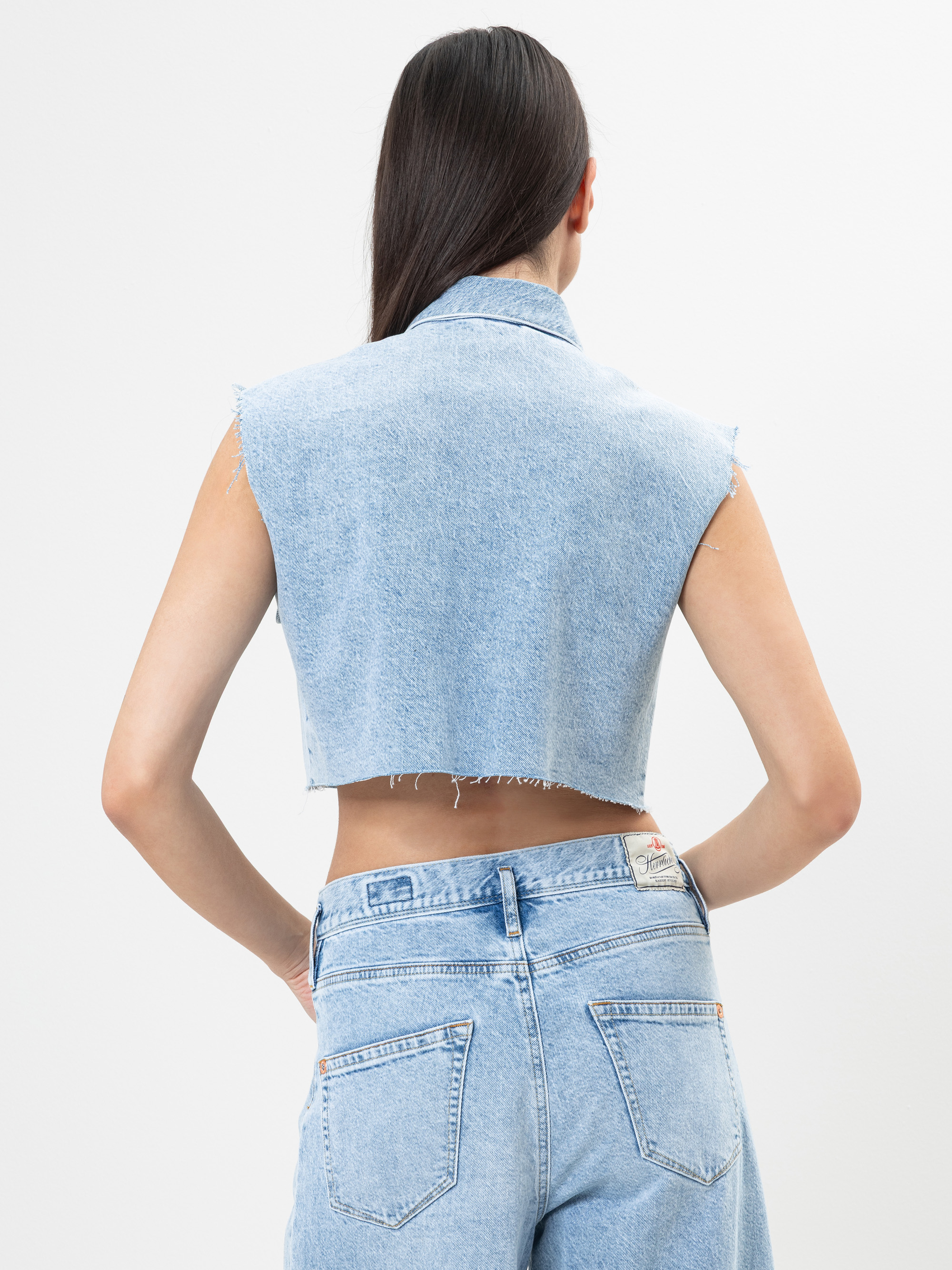 Eine Frau mit langen dunklen Haaren, bekleidet mit einer ärmellosen, hellblauen Jeansweste und hochgeschnittenen hellblauen Jeans, steht mit dem Rücken zur Kamera vor einem schlichten weißen Hintergrund.