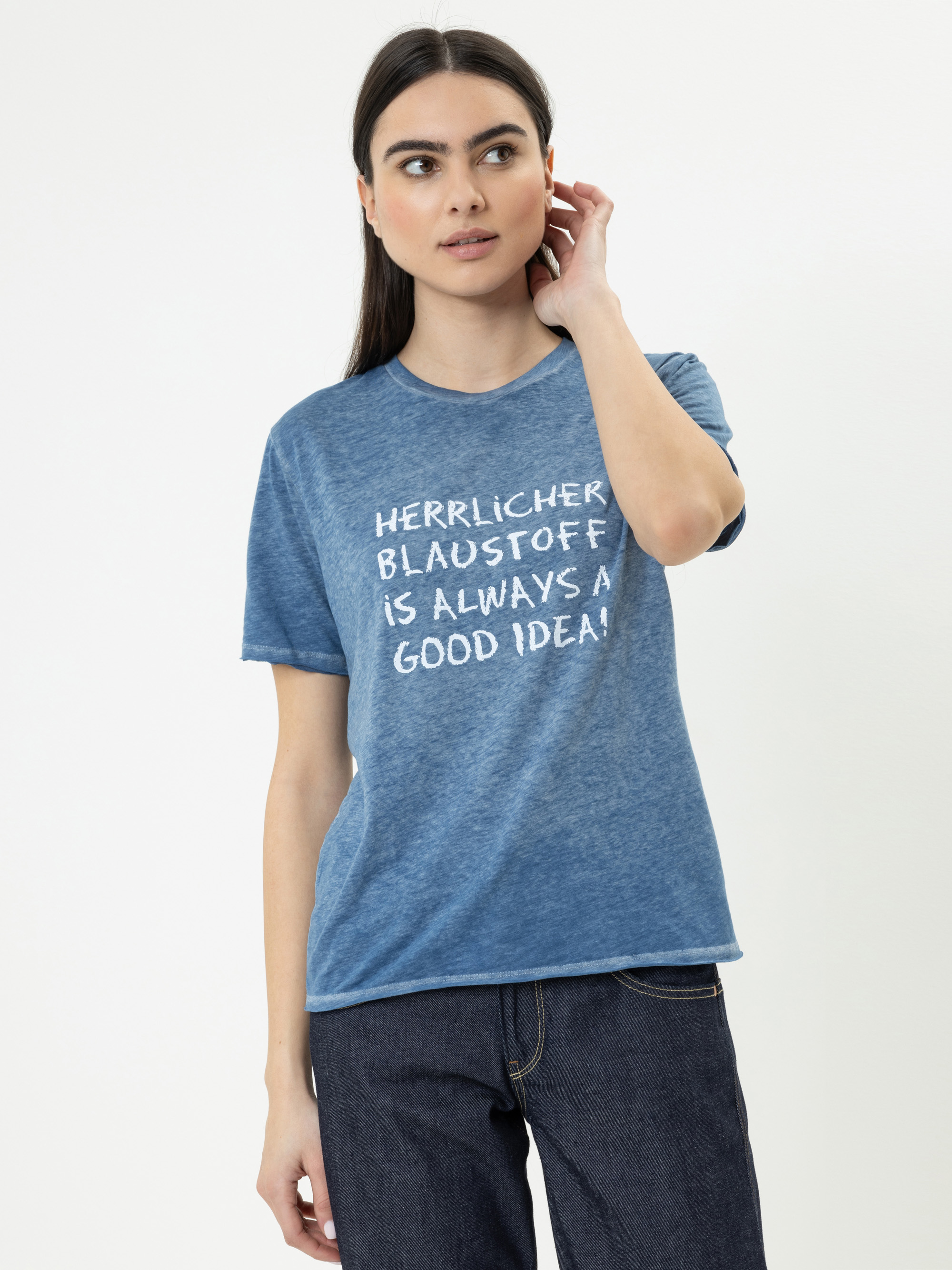 Eine Frau mit langen dunklen Haaren, die ein blaues T-Shirt mit der Aufschrift HERRLICHER BLAUSTOFF IS ALWAYS A GOOD IDEA! und dunkle Jeans trägt, steht vor einem schlichten hellen Hintergrund, berührt ihr Ohr und schaut zur Seite.