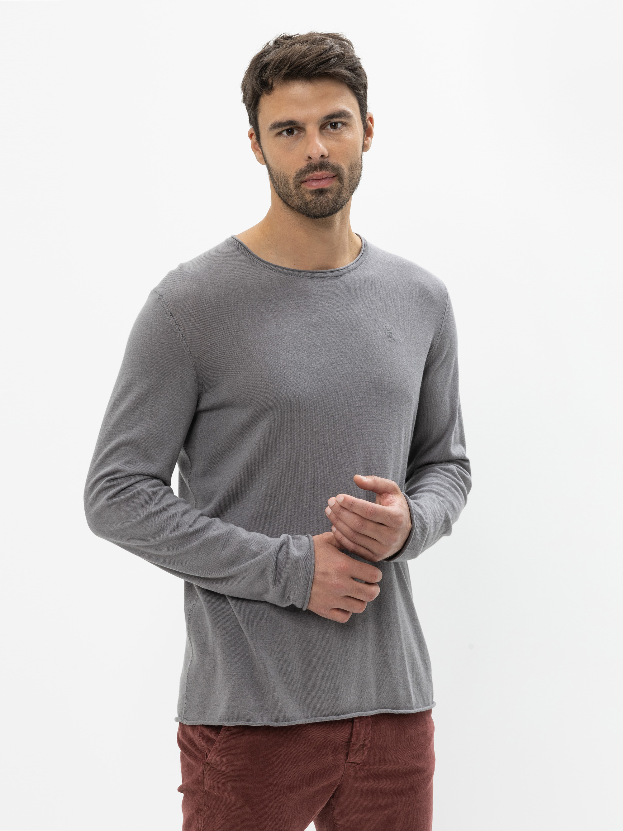 Ein Mann mit kurzen dunklen Haaren und Bart trägt ein langärmeliges, graues Hemd und eine rotbraune Hose. Er steht vor einem schlichten weißen Hintergrund und blickt mit neutralem Blick in die Kamera.