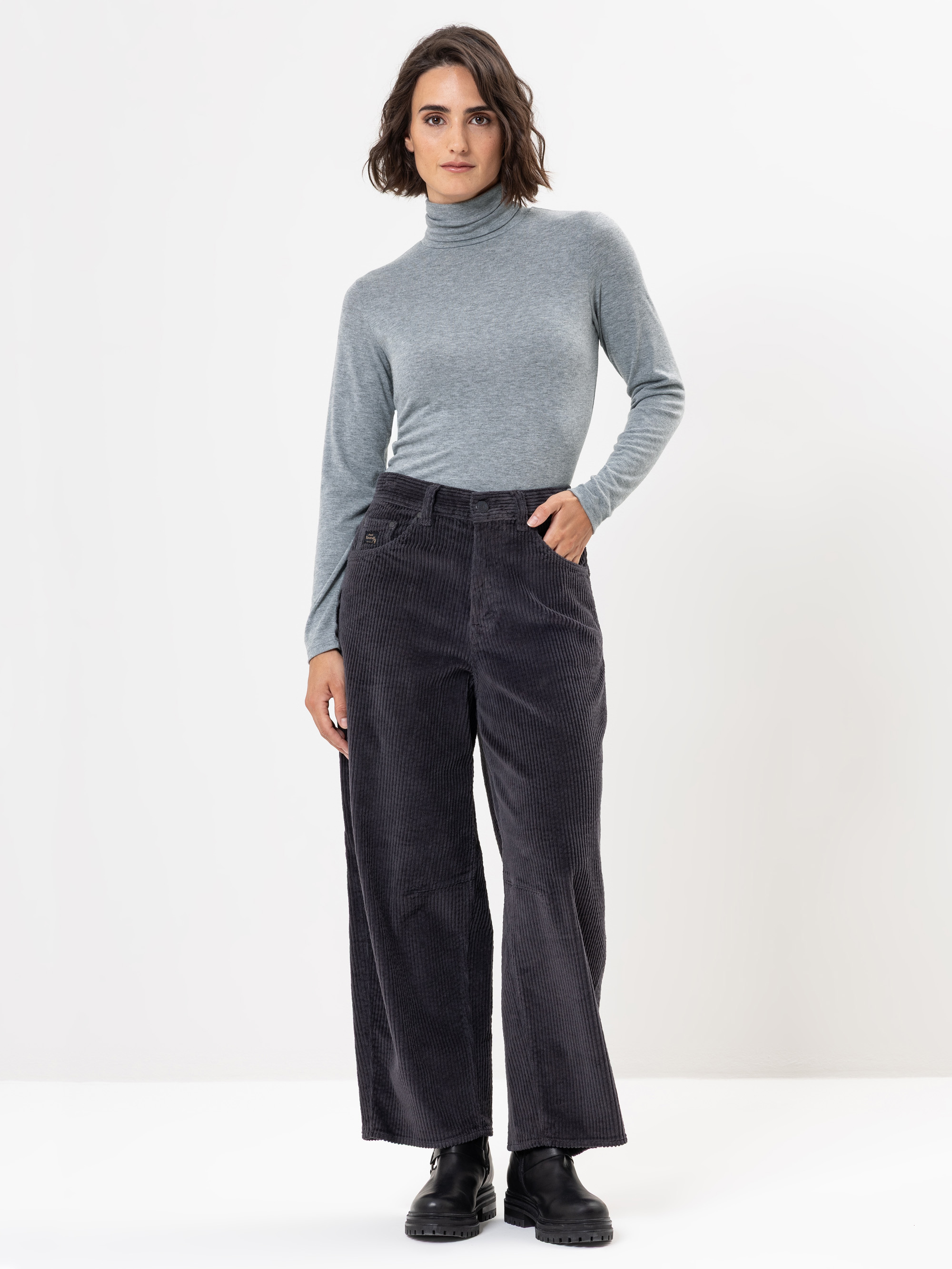 Eine Frau mit schulterlangen braunen Haaren trägt einen hellgrauen Rollkragenpullover, eine dunkle, weit geschnittene Cordhose und schwarze Stiefel und steht selbstbewusst vor einem schlichten weißen Hintergrund.