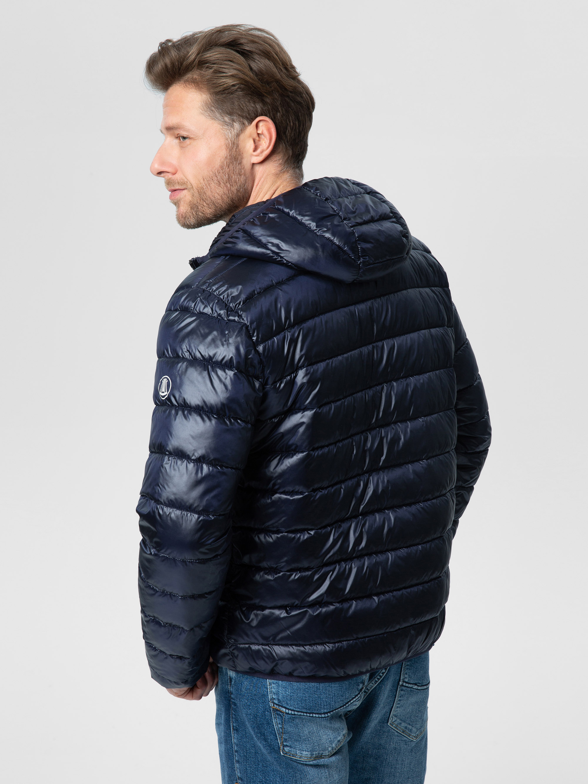 Ein Mann mit kurzen braunen Haaren und einem Bart trägt eine glänzende dunkelblaue Pufferjacke mit Kapuze und blaue Jeans. Er steht mit dem Gesicht zur Seite und schaut über seine linke Schulter vor einem schlichten hellen Hintergrund.