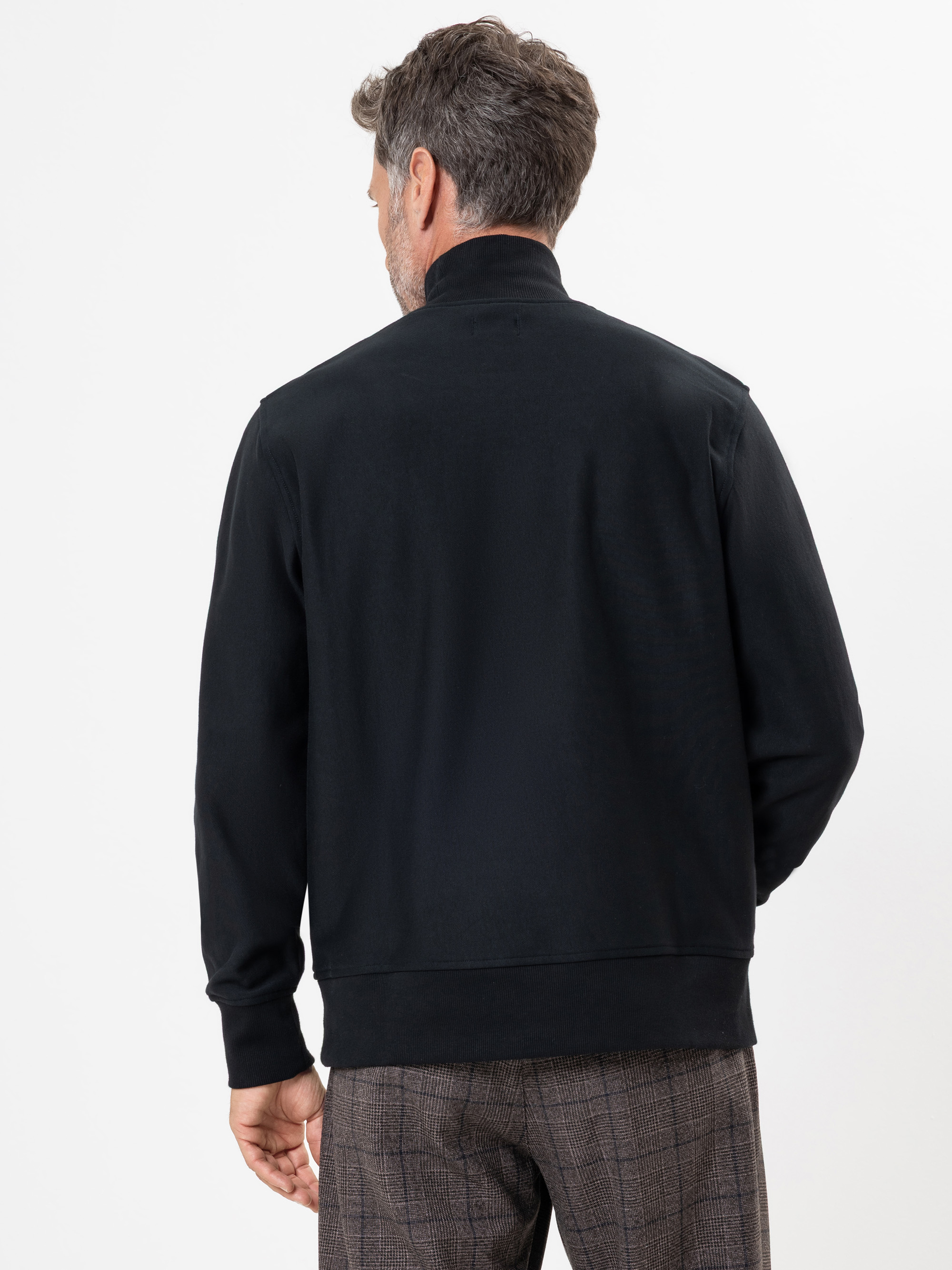 Ein Mann mit kurzem, ergrauendem Haar steht mit dem Rücken zur Kamera, trägt eine schwarze Jacke und eine braun karierte Hose vor einem schlichten weißen Hintergrund.