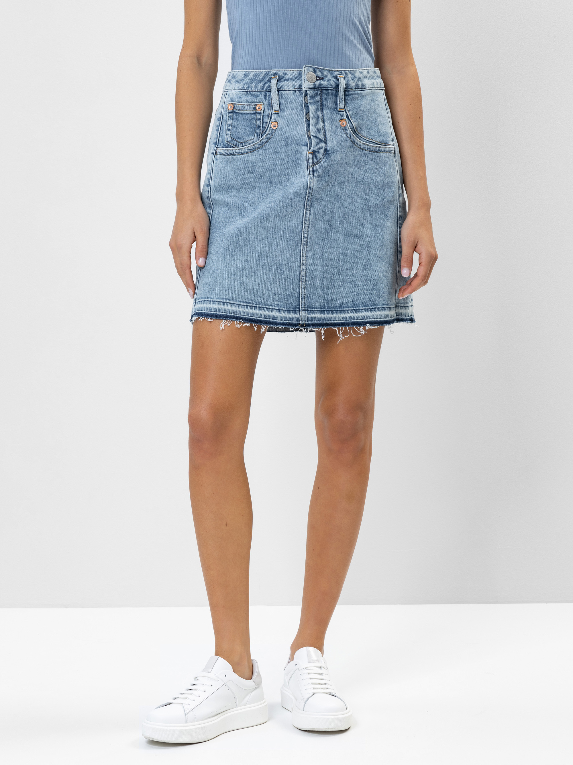 Eine Person, die ein hellblaues geripptes Oberteil, einen hochtaillierten hellblauen Jeansrock mit ausgefranstem Saum und weiße Turnschuhe trägt, steht vor einem schlichten Hintergrund.