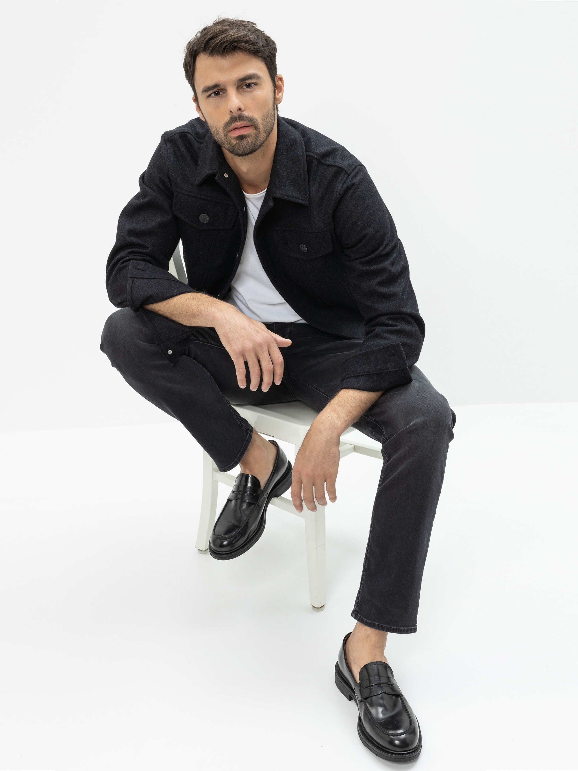 Ein Mann mit kurzen dunklen Haaren und einem Bart sitzt auf einem weißen Stuhl vor einem schlichten weißen Hintergrund. Er trägt eine schwarze Jacke, ein weißes T-Shirt, eine schwarze Hose und schwarze Anzugschuhe und blickt leicht in Richtung der Kam