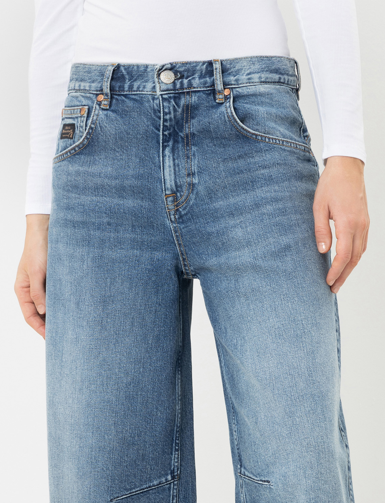 Nahaufnahme des Rückens einer Person, die eine blaue Jeans mit hoher Taille, doppelten Gürtelschlaufen, zwei Gesäßtaschen und einem schwarzen Markenaufnäher aus Leder am Bund trägt. Die Person trägt ein schwarzes Oberteil.