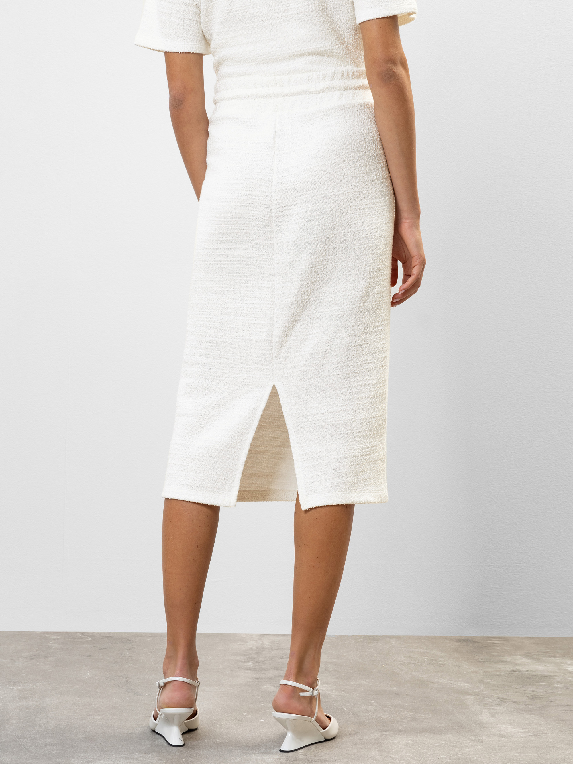 Eine Frau steht auf einem Betonboden und trägt einen weißen, strukturierten Midirock mit Rückenschlitz und weiße, hochhackige Sandalen. Das Foto ist von hinten aufgenommen und zeigt die Passform und Länge des Rocks.