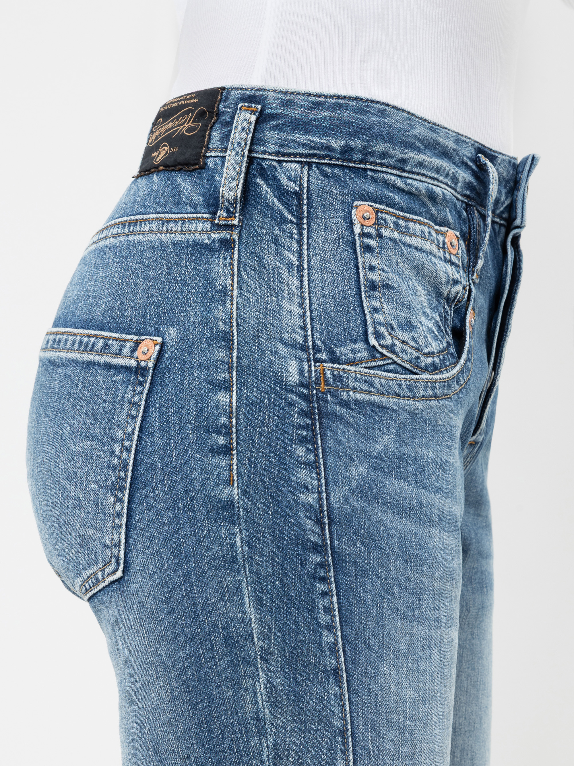 Seitenansicht einer Person, die eine Jeans aus blauem Denim in mittlerer Waschung trägt. Der Fokus liegt auf der Gesäßtasche, der Münztasche und dem schwarzen Lederaufnäher am Bund mit sichtbaren Nahtdetails. Das Model trägt ein schlichtes weißes O