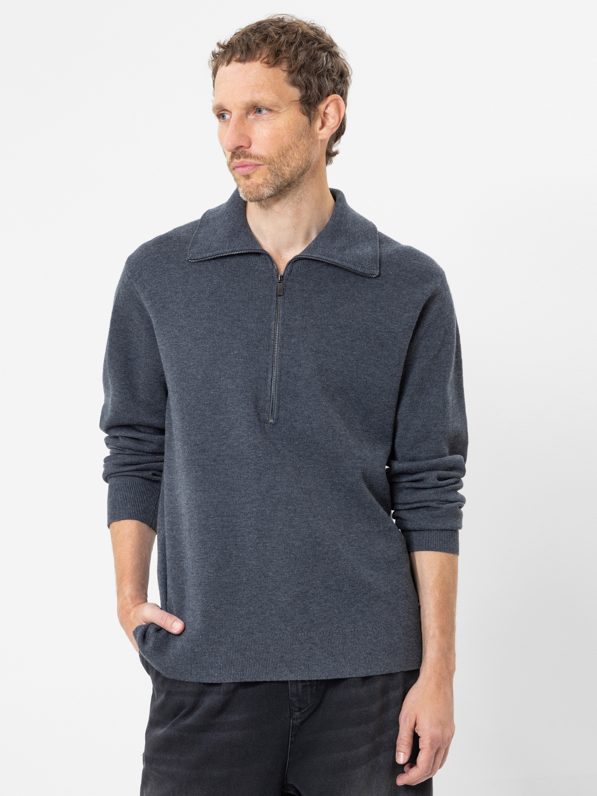Ein Mann mit kurzen braunen Haaren und Bart trägt einen dunkelgrauen Pullover mit Reißverschluss am Kragen und eine schwarze Hose; er steht vor einem schlichten weißen Hintergrund und hat eine Hand in der Tasche.