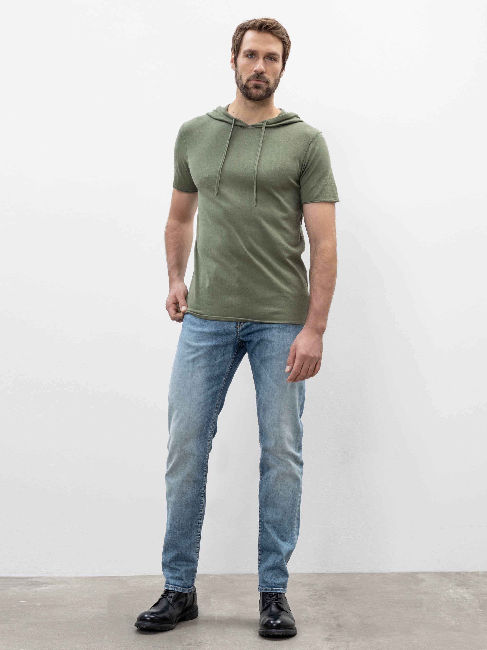 Ein Mann steht vor einer schlichten weißen Wand und trägt einen kurzärmeligen olivgrünen Kapuzenpullover, hellblaue Jeans und schwarze Schuhe. Er hat kurzes braunes Haar und einen Bart, einen neutralen Gesichtsausdruck und eine entspannte Körperhaltu