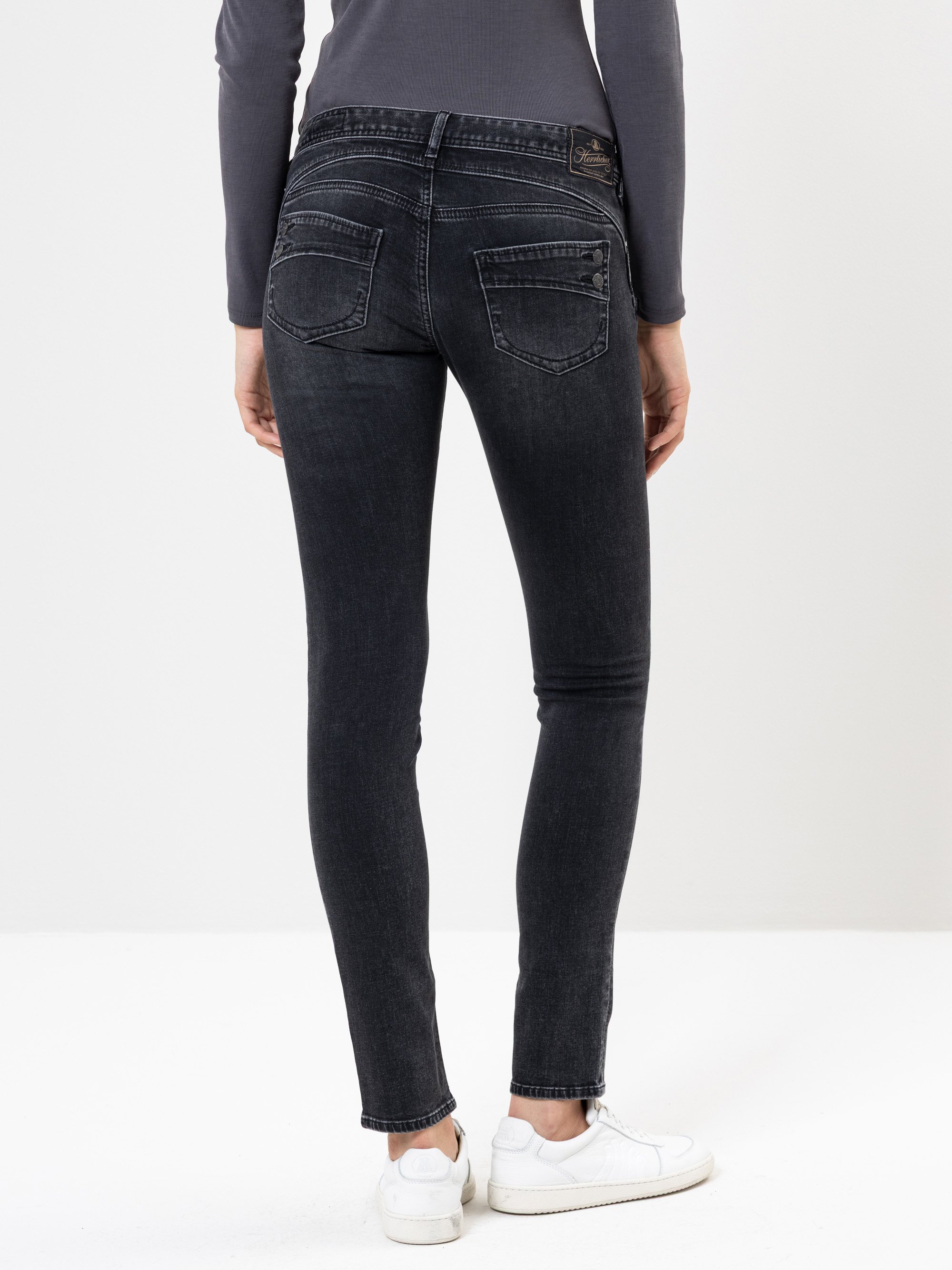 Eine Person, die ein langärmeliges graues Oberteil, eine schwarze Skinny-Jeans und weiße Turnschuhe trägt, steht mit dem Gesicht nach hinten und zeigt die Rückenansicht der Jeans vor einem schlichten weißen Hintergrund.