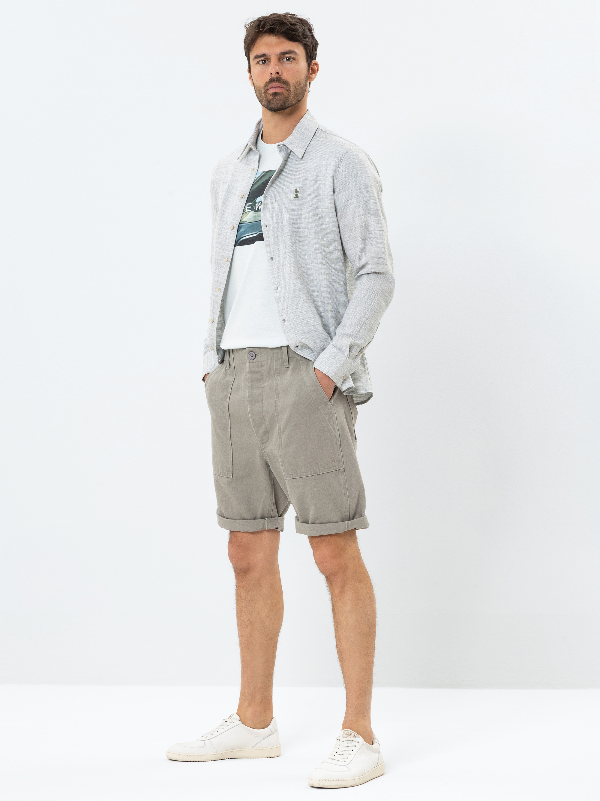 Ein Mann steht vor einem schlichten weißen Hintergrund und trägt ein hellgraues Button-up-Hemd über einem bedruckten weißen T-Shirt, beigefarbene Shorts mit aufgerollten Manschetten und weiße Turnschuhe. Er hat die Hände in den Taschen und blickt le