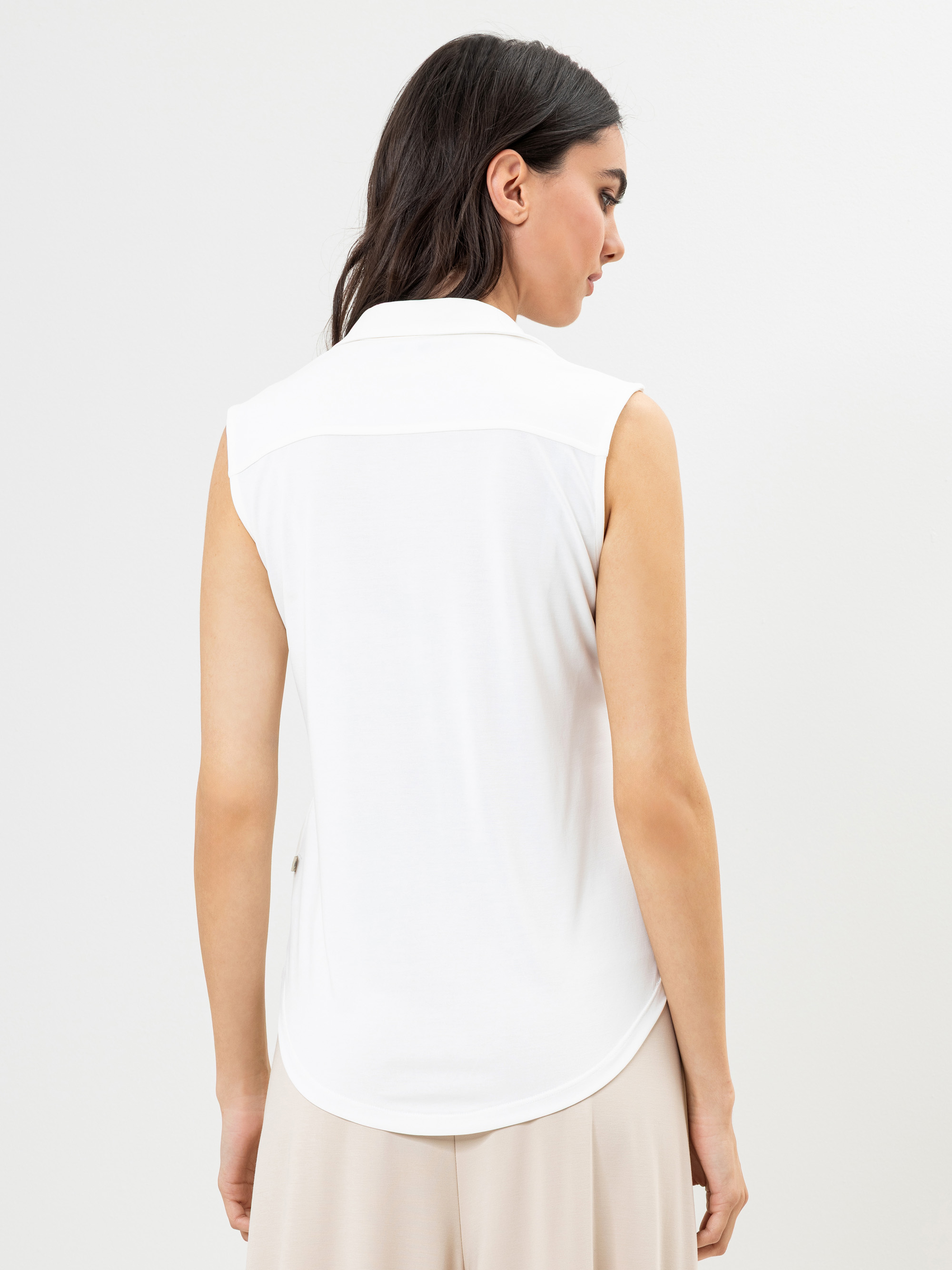 Man sieht eine Frau mit langen dunklen Haaren von hinten, die ein ärmelloses weißes Hemd mit Kragen und eine hellbeige Hose trägt und vor einem schlichten weißen Hintergrund steht.