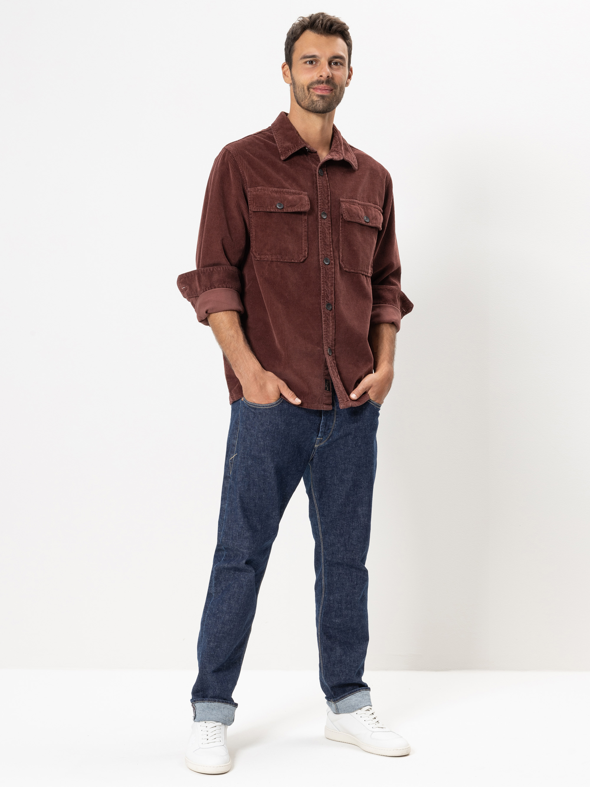 Ein Mann steht vor einem weißen Hintergrund und trägt ein kastanienbraunes Button-up-Hemd mit Brusttaschen, blaue Jeans und weiße Turnschuhe. Seine Hände stecken in den Taschen und er lächelt leicht.
