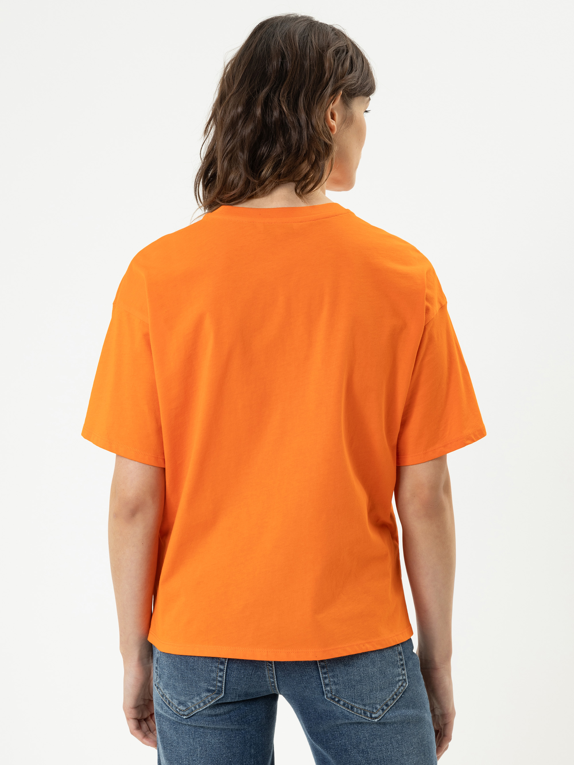 Eine Person mit gewelltem braunem Haar wird von hinten gezeigt, die ein orangefarbenes kurzärmeliges T-Shirt und blaue Jeans vor einem einfarbig weißen Hintergrund trägt.